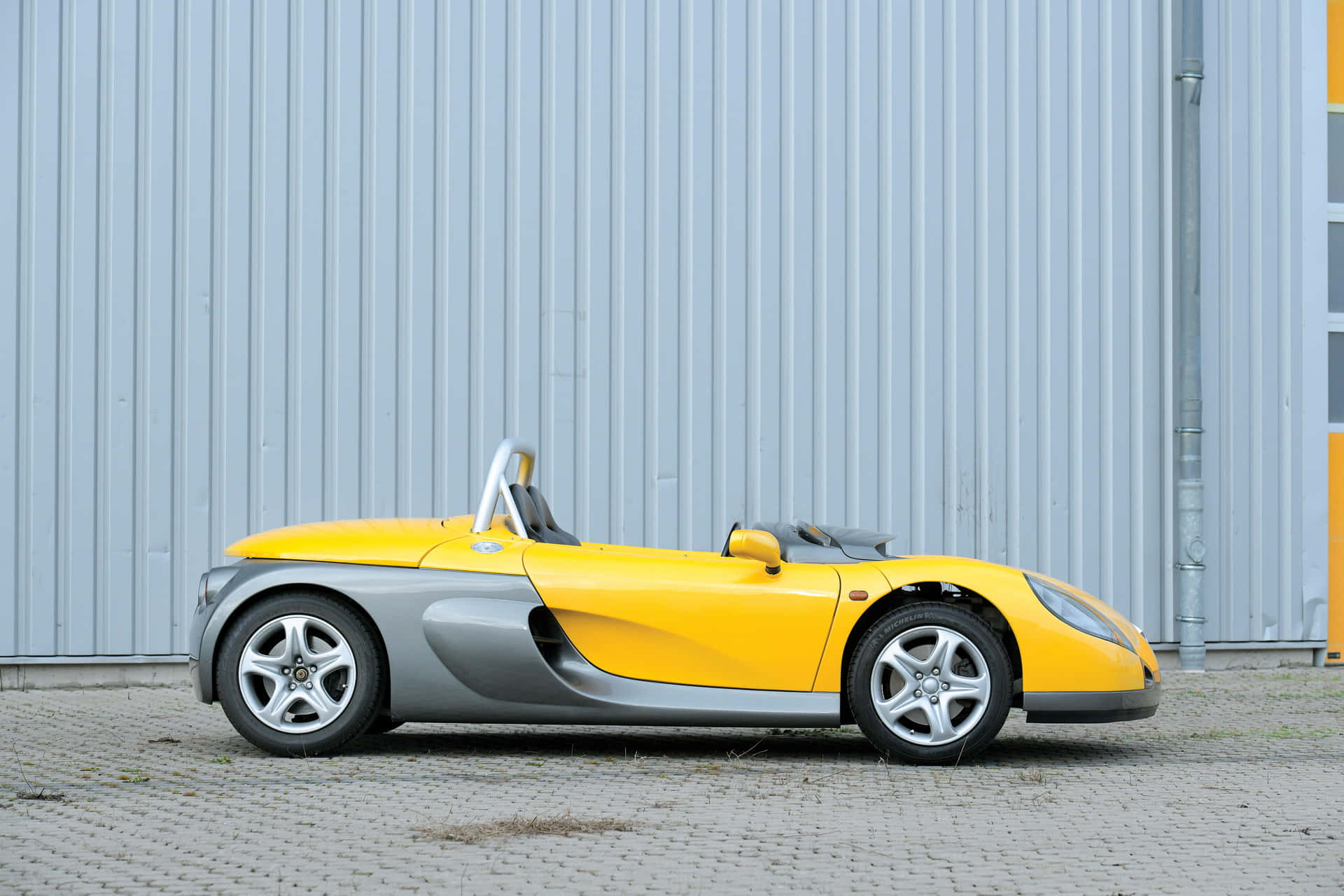Renault Sport Spider 桌布 壁紙 — 一輛鮮黃色和銀色的跑車停泊在鋪砌路面上，靠著波紋金屬牆。