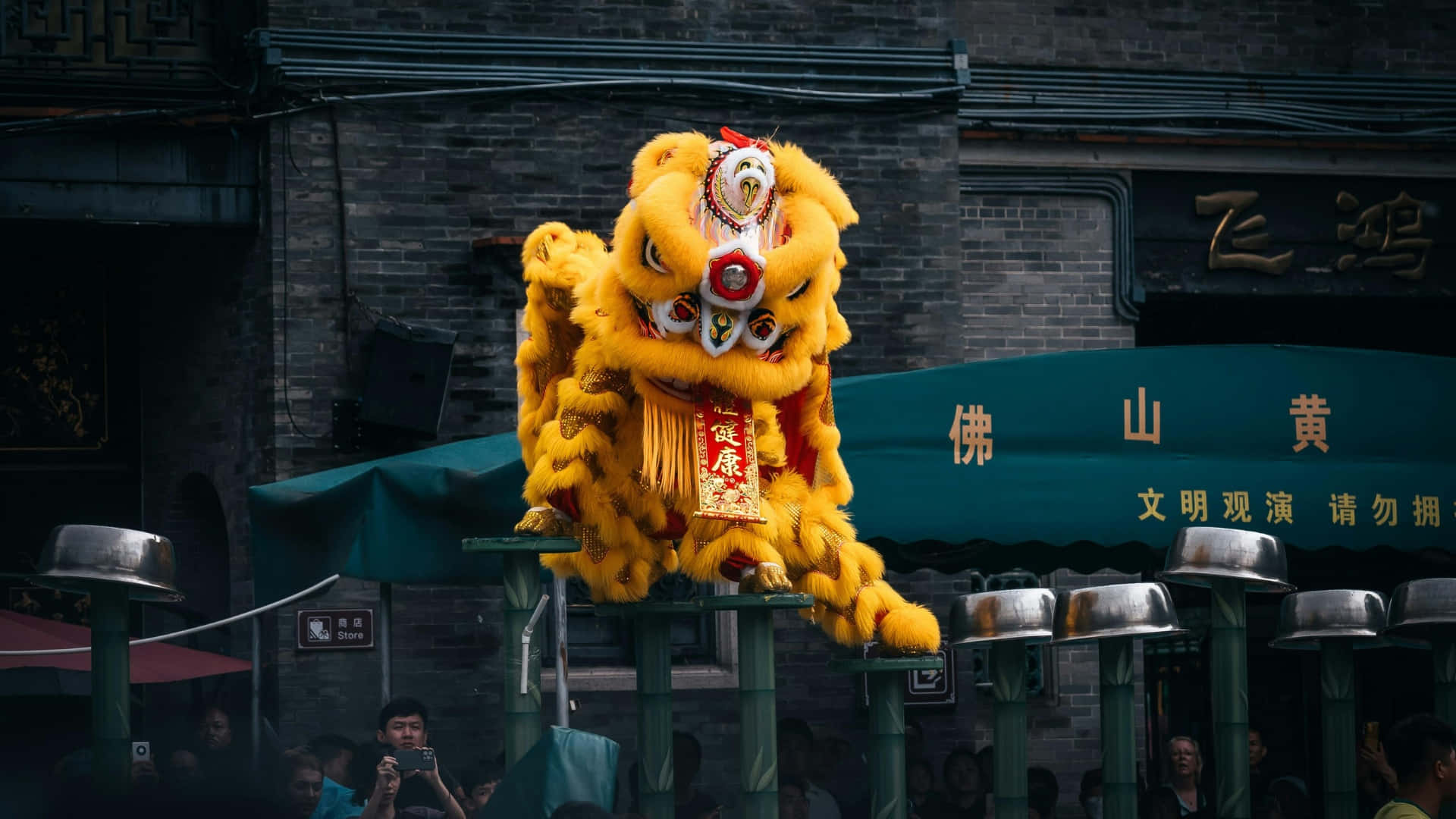 壁紙 — 鮮黃色嘅舞獅服裝喺有中文字嘅深色磚建築前面踩高蹺表演。
