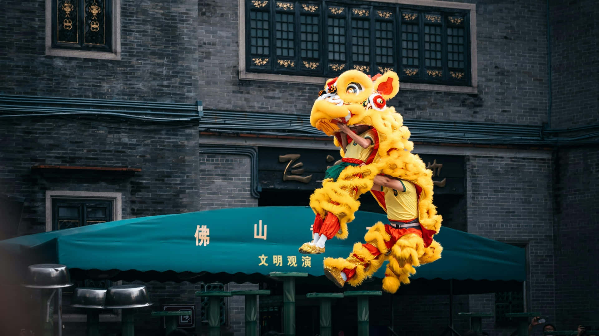  壁紙 — 充滿活力的黃色舞獅表演者在歷史悠久的灰色磚建築外牆前動態跳躍。
