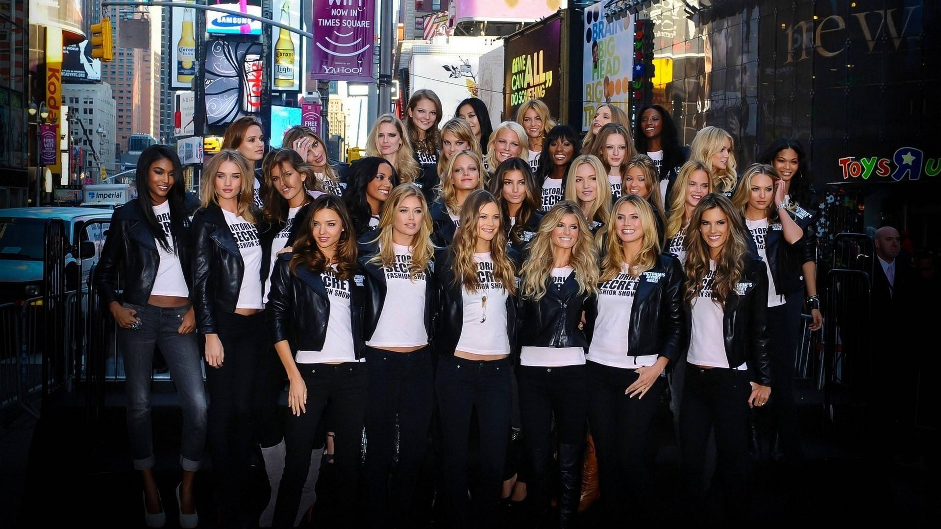 Victoria's Secret 天使於時代廣場 壁紙 — 一群穿著黑色皮褸和白色T恤的女性在時代廣場明亮的霓虹燈街道上一起擺姿勢。