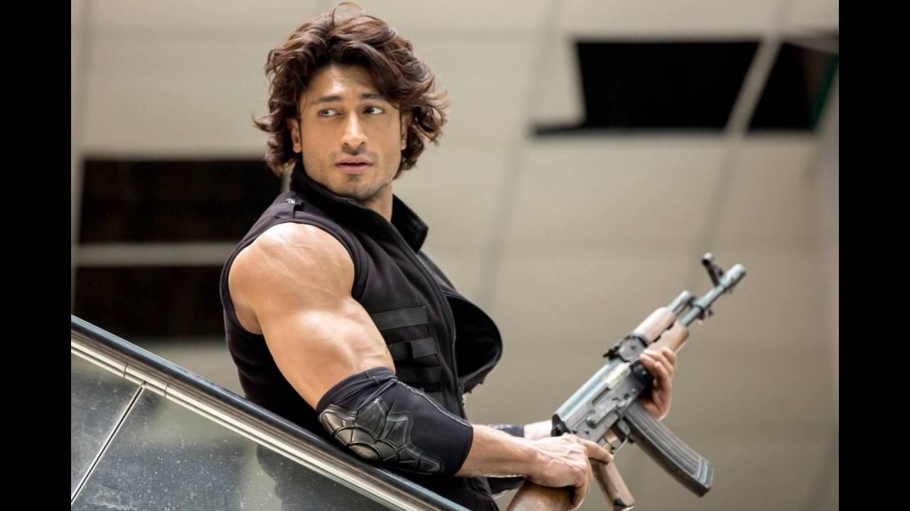 Vidyut Jamwal 手持步槍 壁紙 — 一個肌肉發達、波浪頭髮的男人在現代都市環境中靠著一輛車，手持突擊步槍。