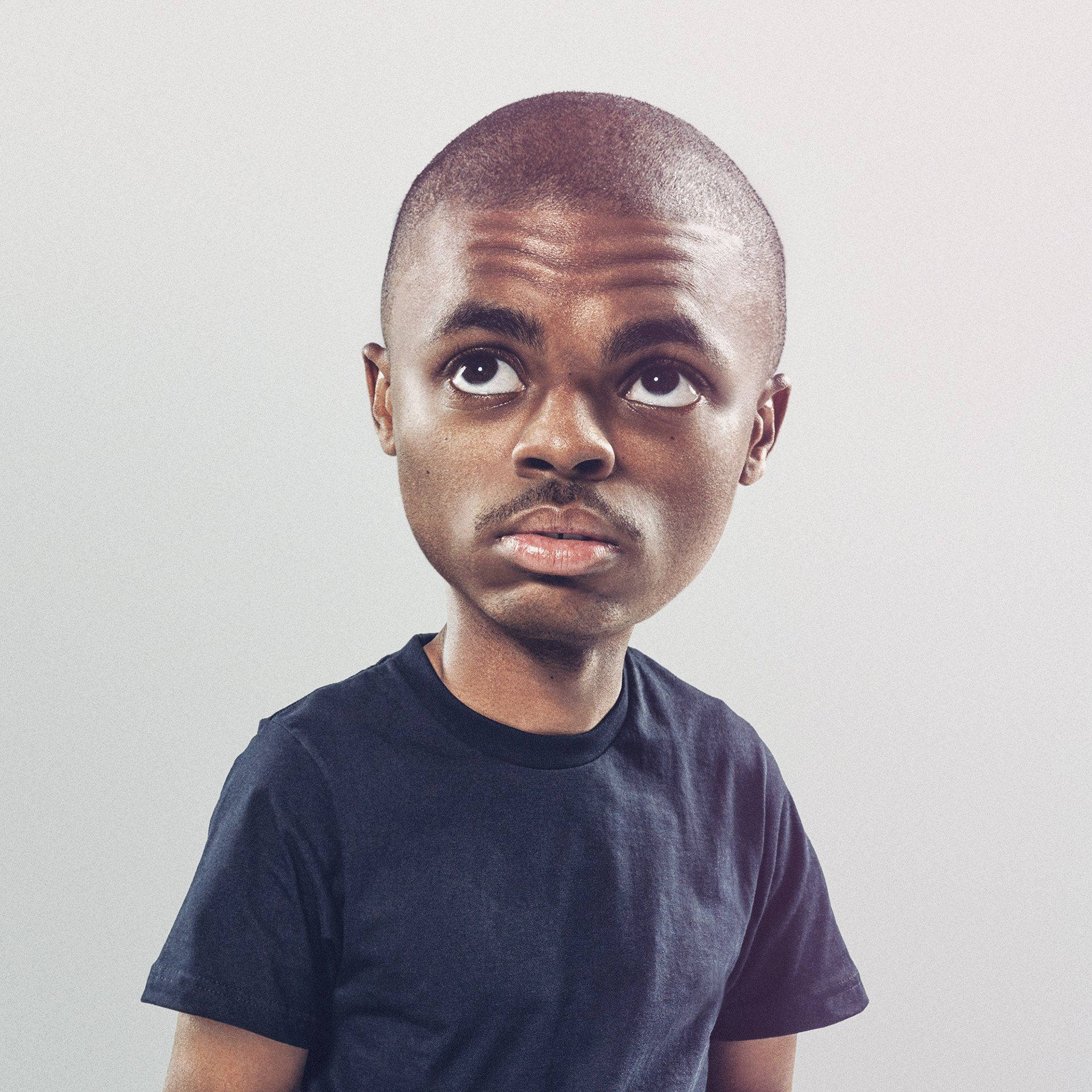 大頭Vince Staples 壁紙 — 一個剃光頭、身穿深色T恤的年輕男子，在純灰色背景前睜大眼睛向上凝視。