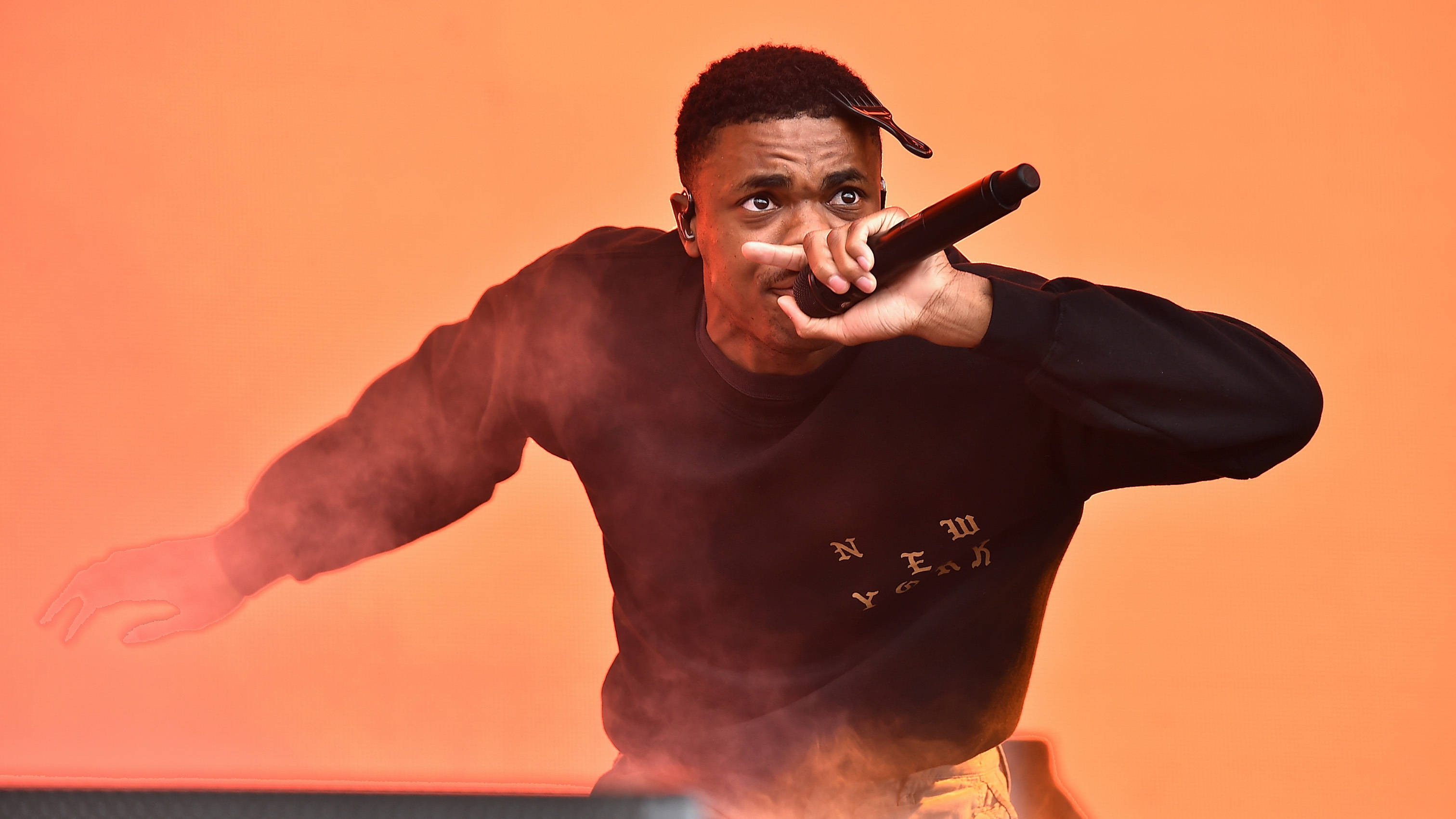美學 Vince Staples 壁紙 — 一位身穿黑色運動衫的饒舌歌手在充滿活力的橙色煙霧背景下對著麥克風熱情表演。