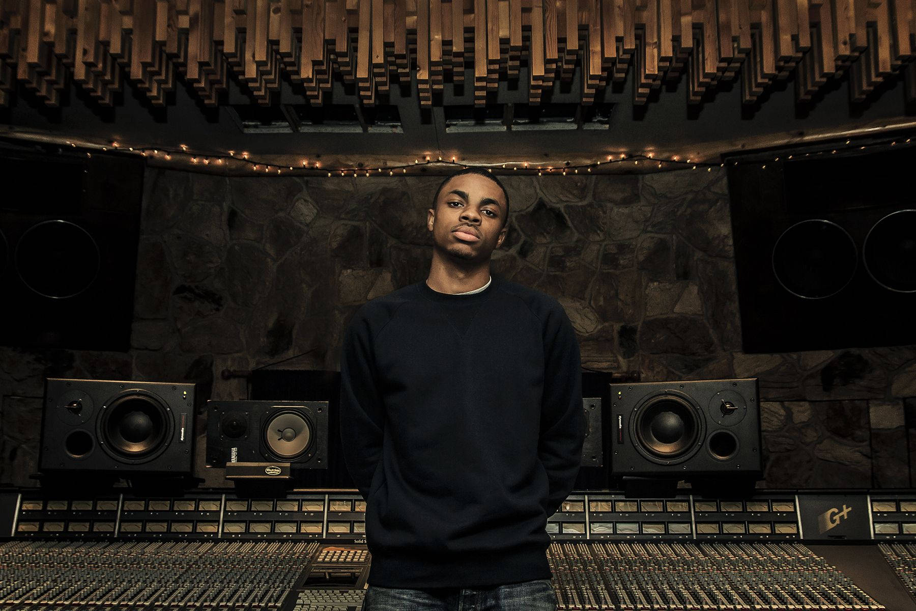 Vince Staples 在錄音室 壁紙 — 一位年輕男子自信地站在燈光昏暗的專業錄音室中，周圍環繞著揚聲器和混音台。