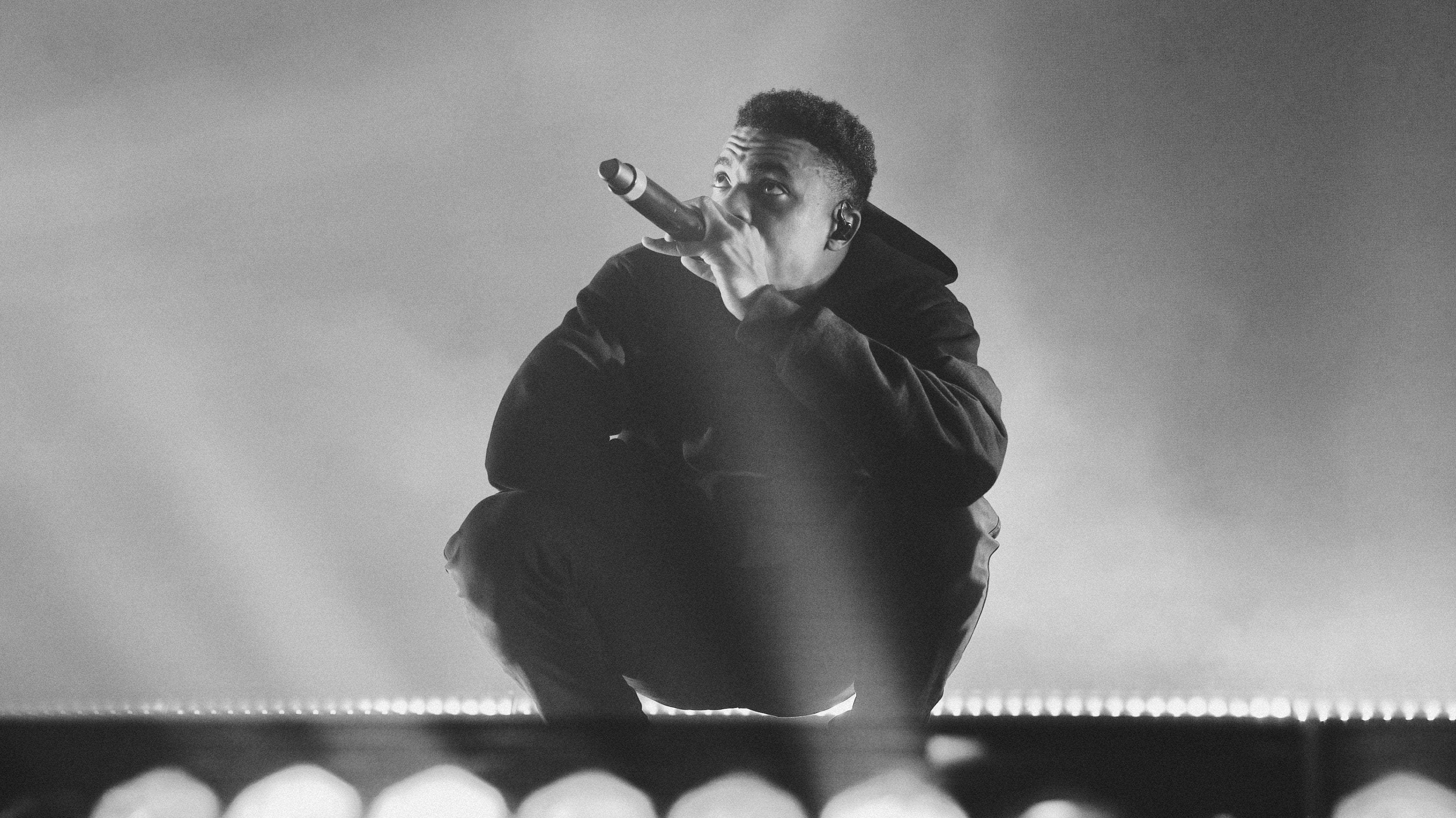 Vince Staples 演唱會現場 壁紙 — 一張黑白照片，一名說唱歌手在戲劇性舞台燈光下蹲在舞台上，手持麥克風。