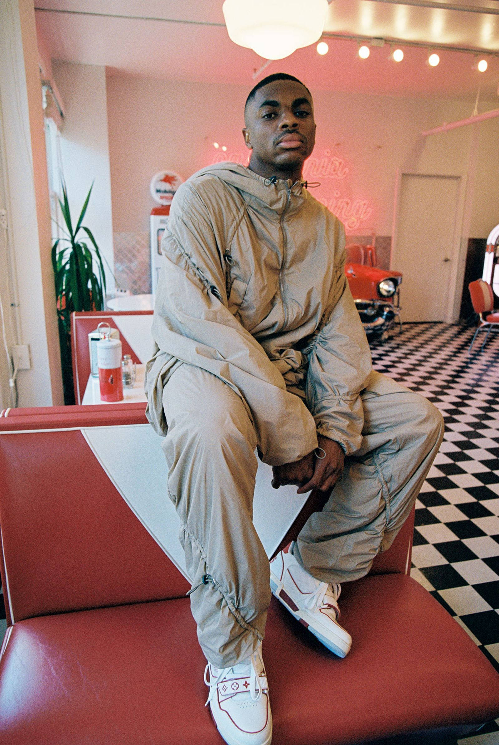 Vince Staples 精緻攝影寫真 壁紙 — 一位身穿米色運動服的年輕男子自信地坐在紅色餐廳卡座上，地板是棋盤格紋，霓虹燈牌發出粉紅光芒。