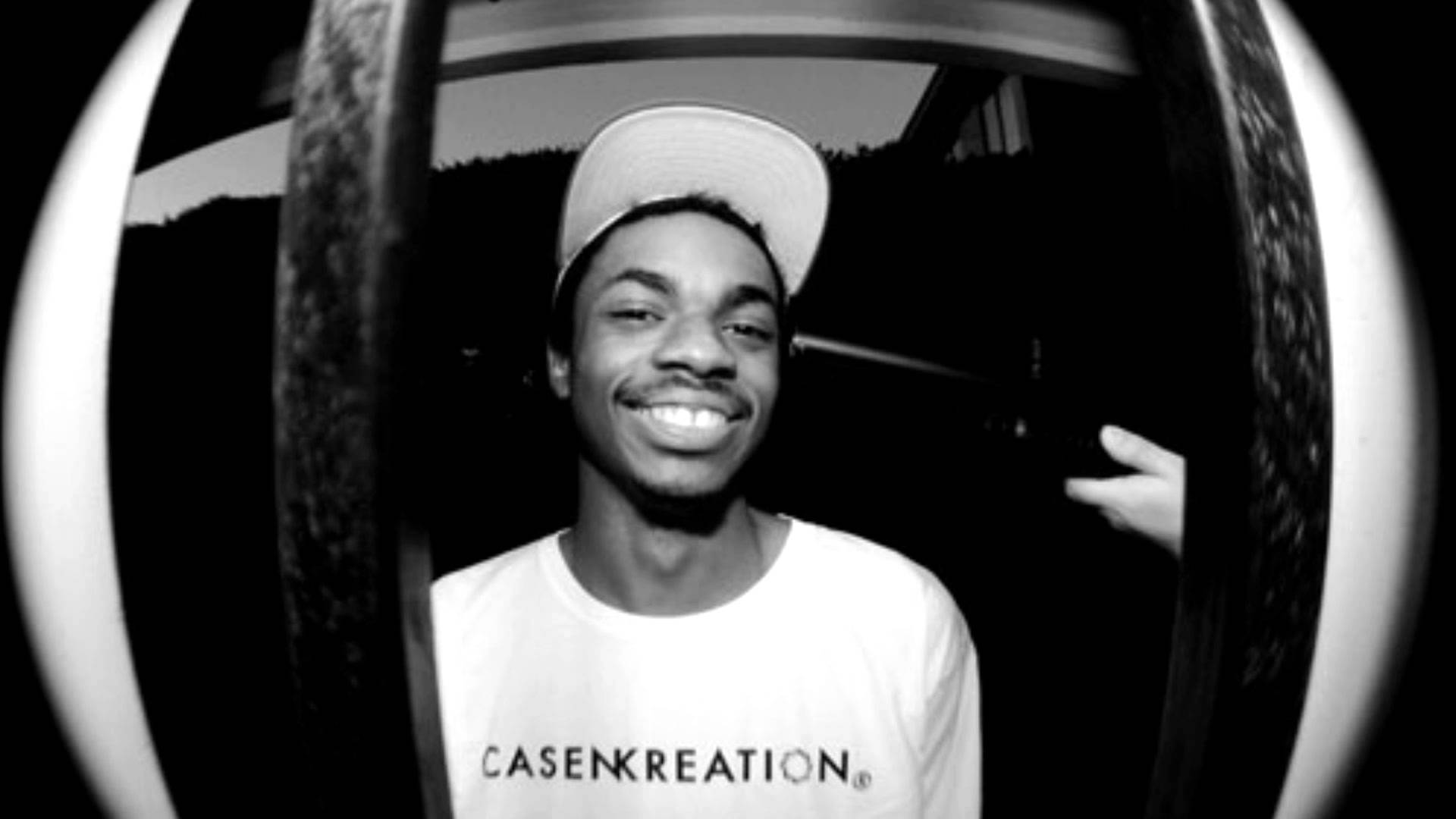 Vince Staples 魚眼鏡頭 壁紙 — 一位微笑的年輕男子戴著白色帽子穿著T恤，透過車門上的魚眼鏡頭反射觀看。