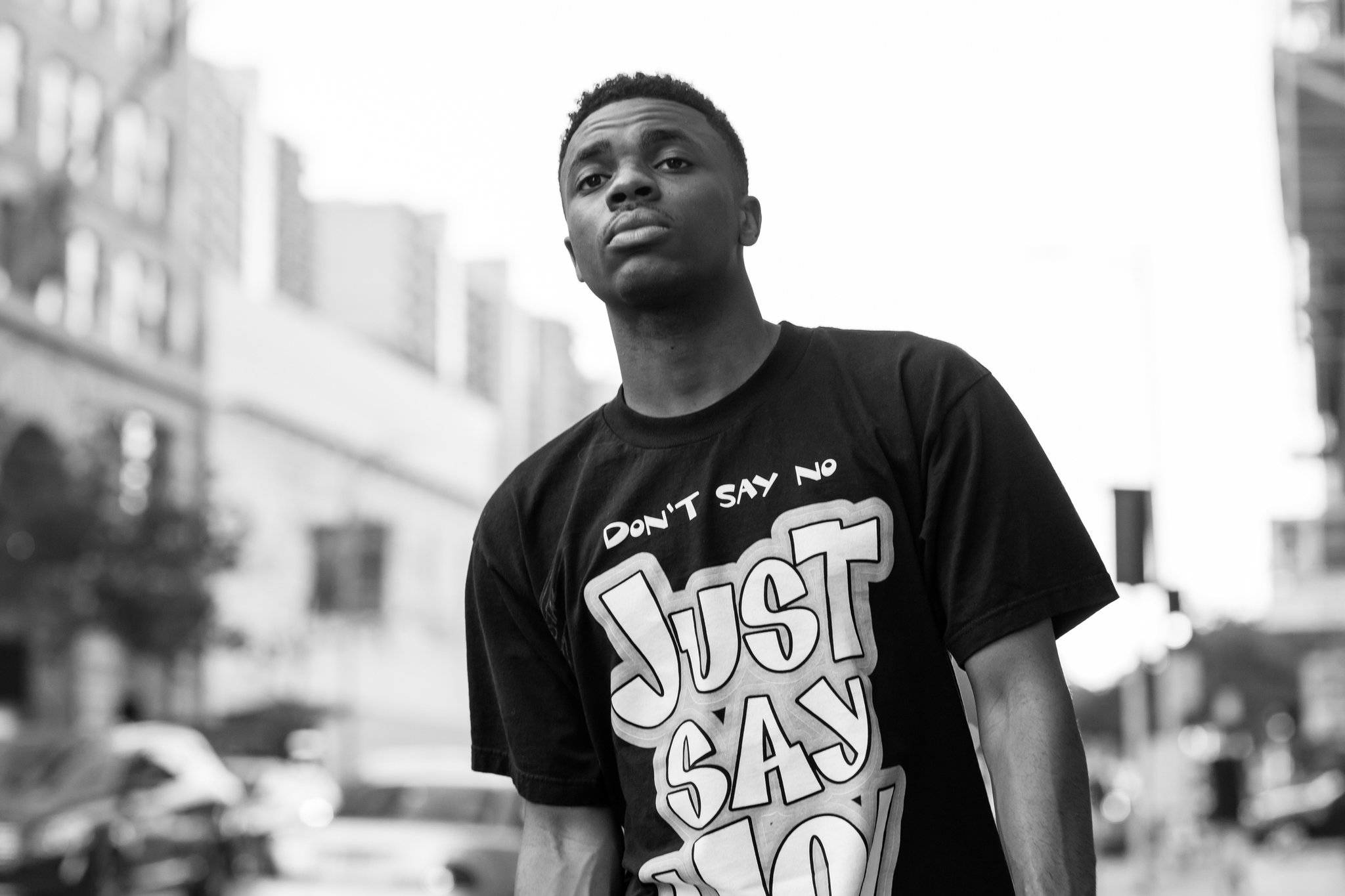 Vince Staples 黑白街頭風 壁紙 — 一位穿著大膽T恤的年輕男子自信地站在城市街道上，黑白畫面，身後建築物模糊。