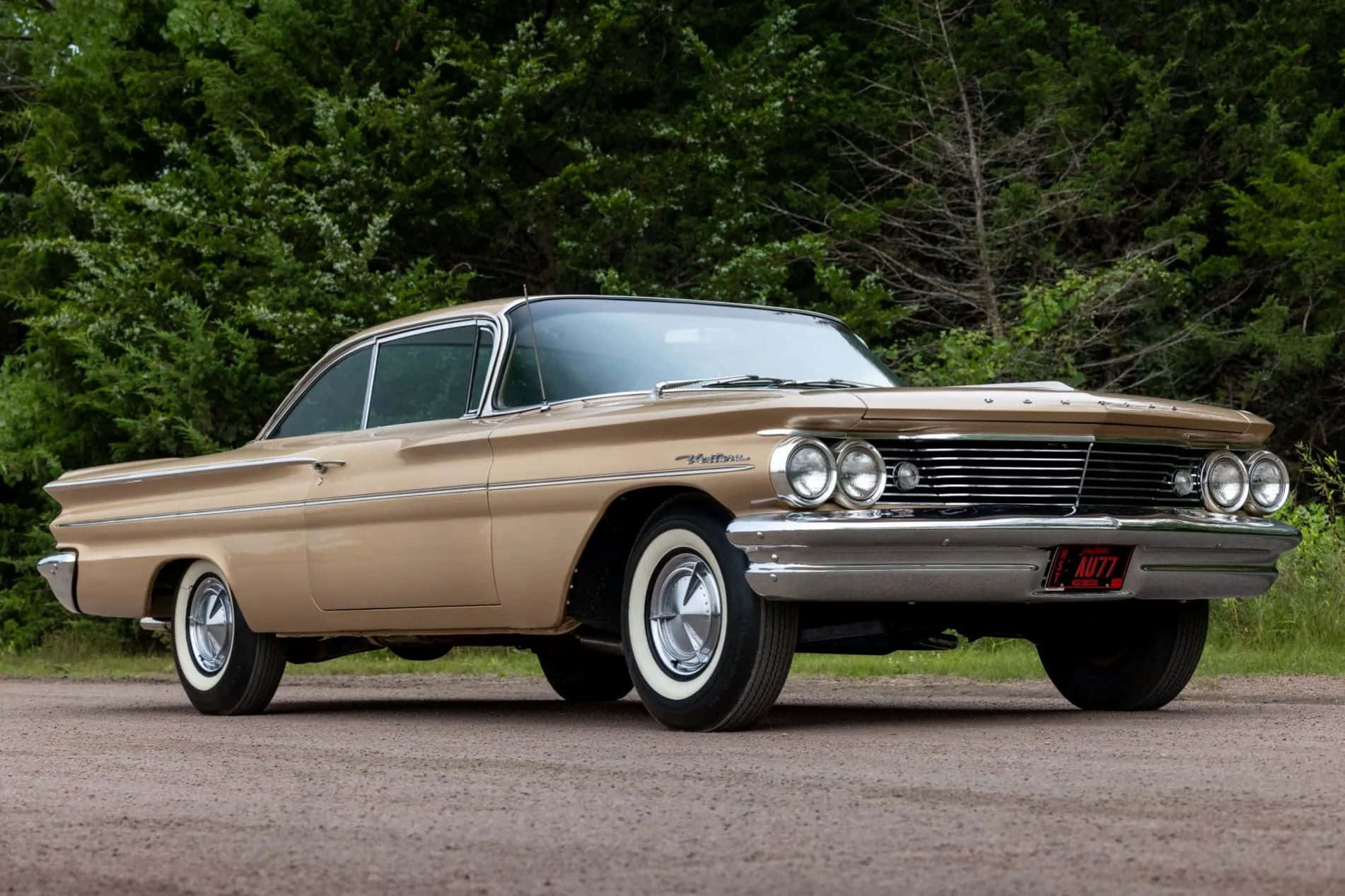  壁紙 — 一輛復古米色1960年雪佛蘭Bel Air，帶有鍍鉻裝飾和白牆輪胎，停在碎石路上，旁邊是茂密的綠樹。