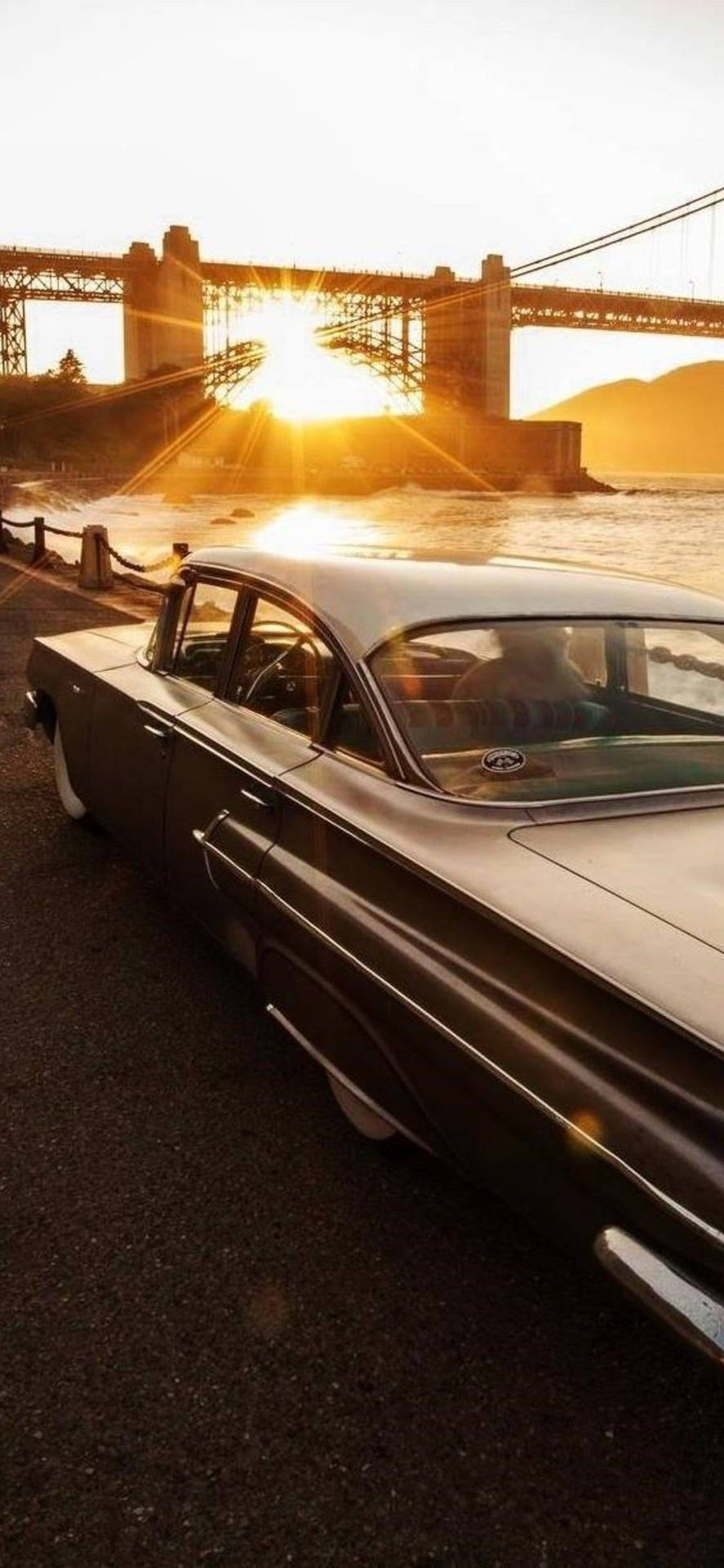 垂直畫面的復古凱迪拉克日落 壁紙 — 一輛復古汽車在金色夕陽下閃閃發光，停在水邊的橋旁。