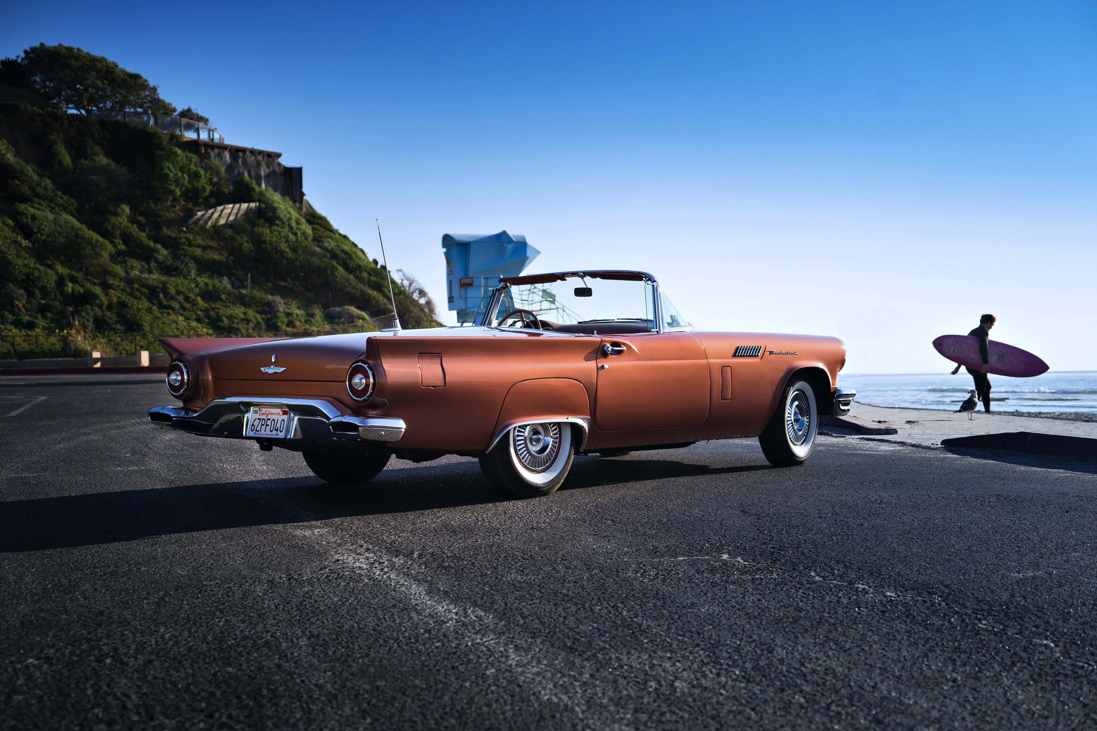 Ford Thunderbird 桌布 壁紙 — 一輛復古銅色開篷車停泊在海岸公路上，一名衝浪者在晴朗的藍天下走向海洋。