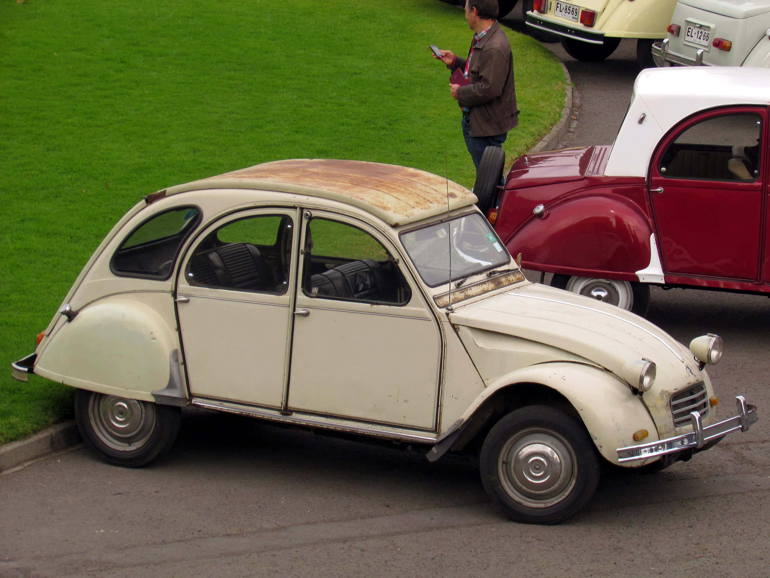 Citroen Ax 桌布 壁紙 — 一架復古奶油色雪鐵龍2CV，帶有生鏽車頂，停泊在街上，旁邊有一輛紅白經典汽車。