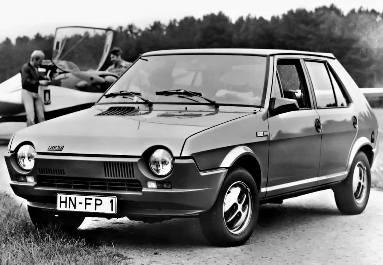 Fiat Ritmo 桌布 壁紙 — 一輛復古菲亞特126p汽車停泊在泥路上，背景有兩名男子靠近一架小型飛機。