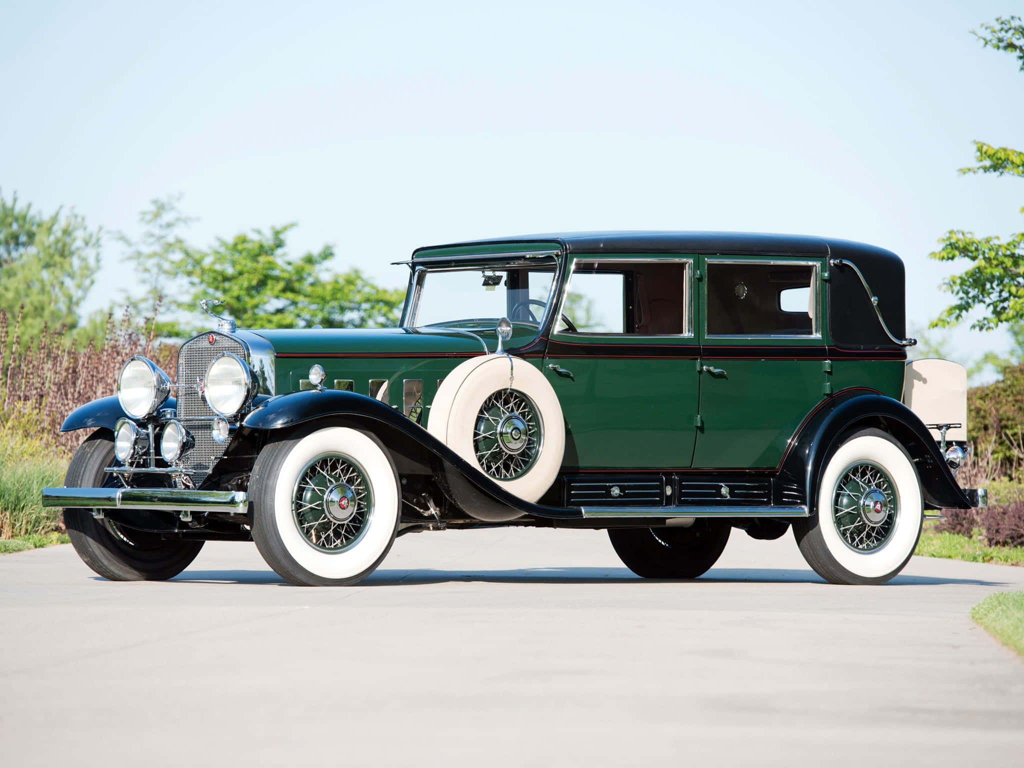 Cadillac Fleetwood 桌布 壁紙 — 一輛復古綠色豪華車，配備白色側壁輪胎和鍍鉻裝飾，停在鋪砌的道路上，周圍綠樹成蔭，天空晴朗。