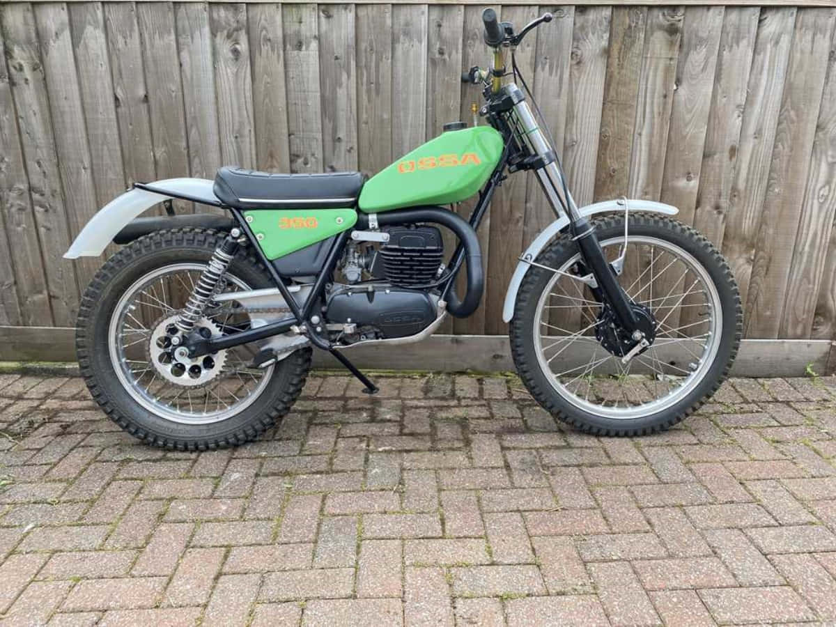 Ossa Motorcycle 桌布 壁紙 — 一架復古綠色USAA 350cc越野電單車停泊在磚砌庭院，背景是風化的木柵欄。