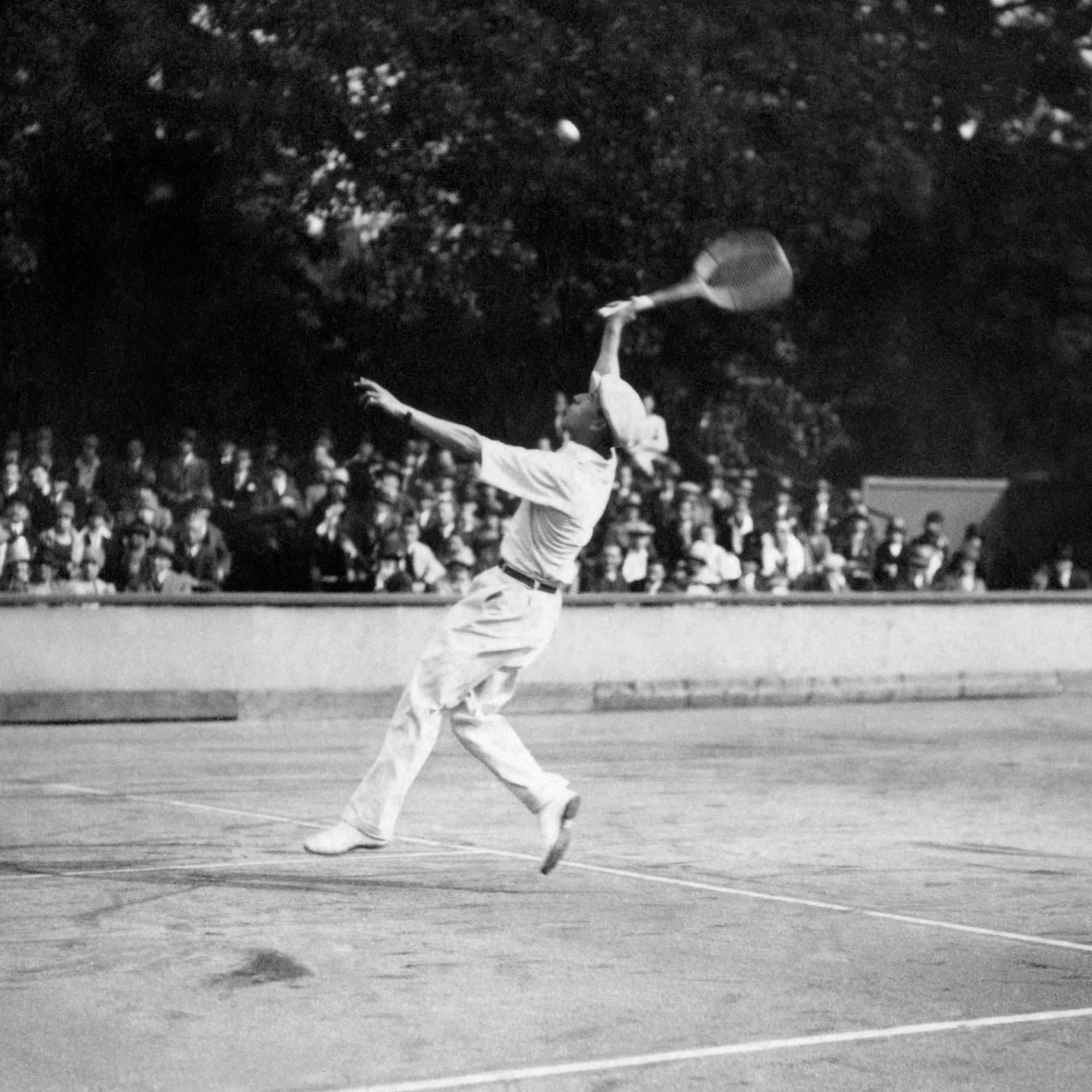 René Lacoste 注視球拍 壁紙 — 一張復古黑白照片捕捉到一名網球選手在半空中執行強力過頭扣殺，在陽光明媚的戶外球場上，有觀眾觀看。