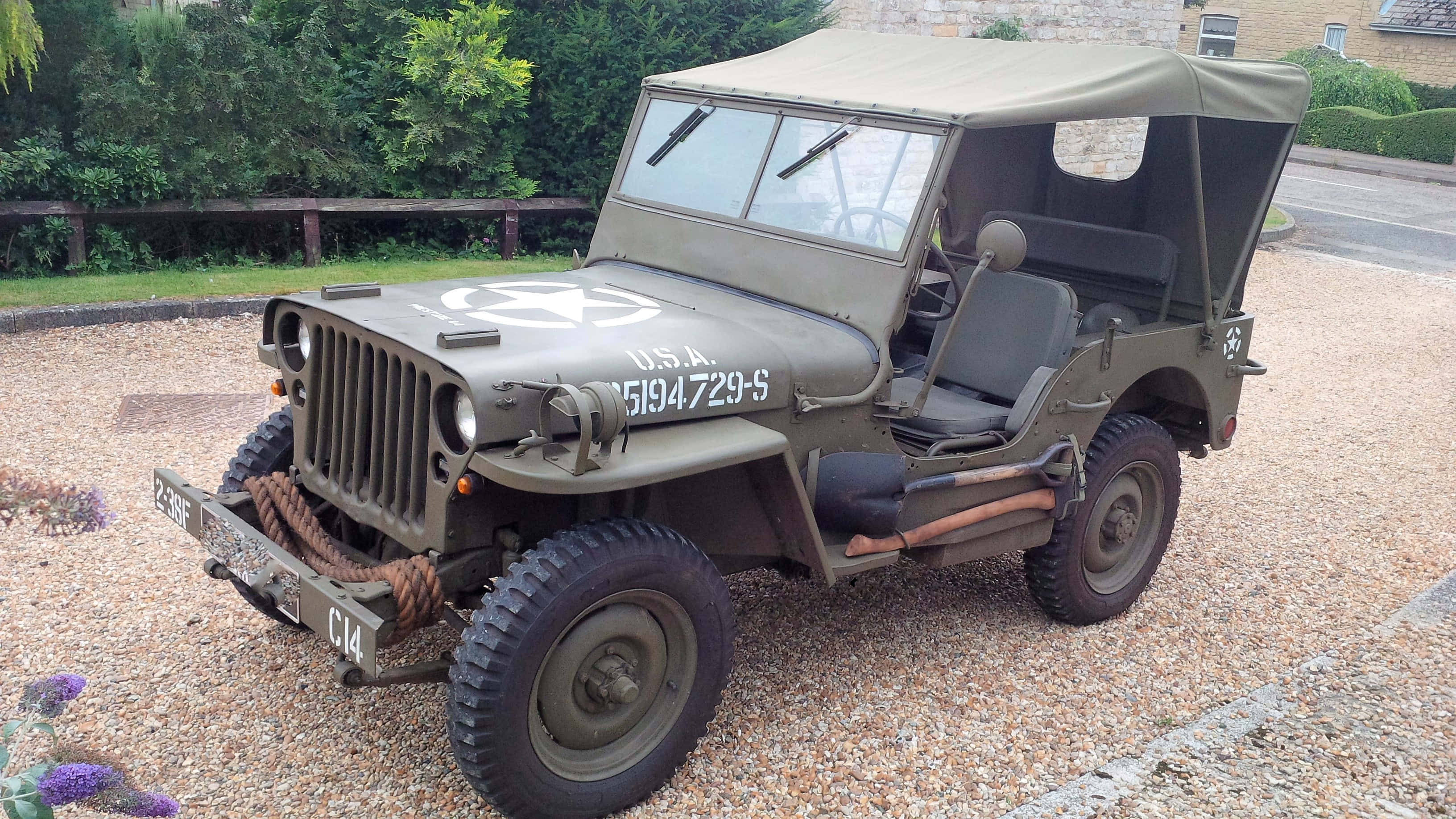 Jeep Willys 桌布 壁紙 — 一輛復古橄欖綠軍用吉普車，帶帆布頂篷，停在碎石上，旁邊有綠植和石頭建築。