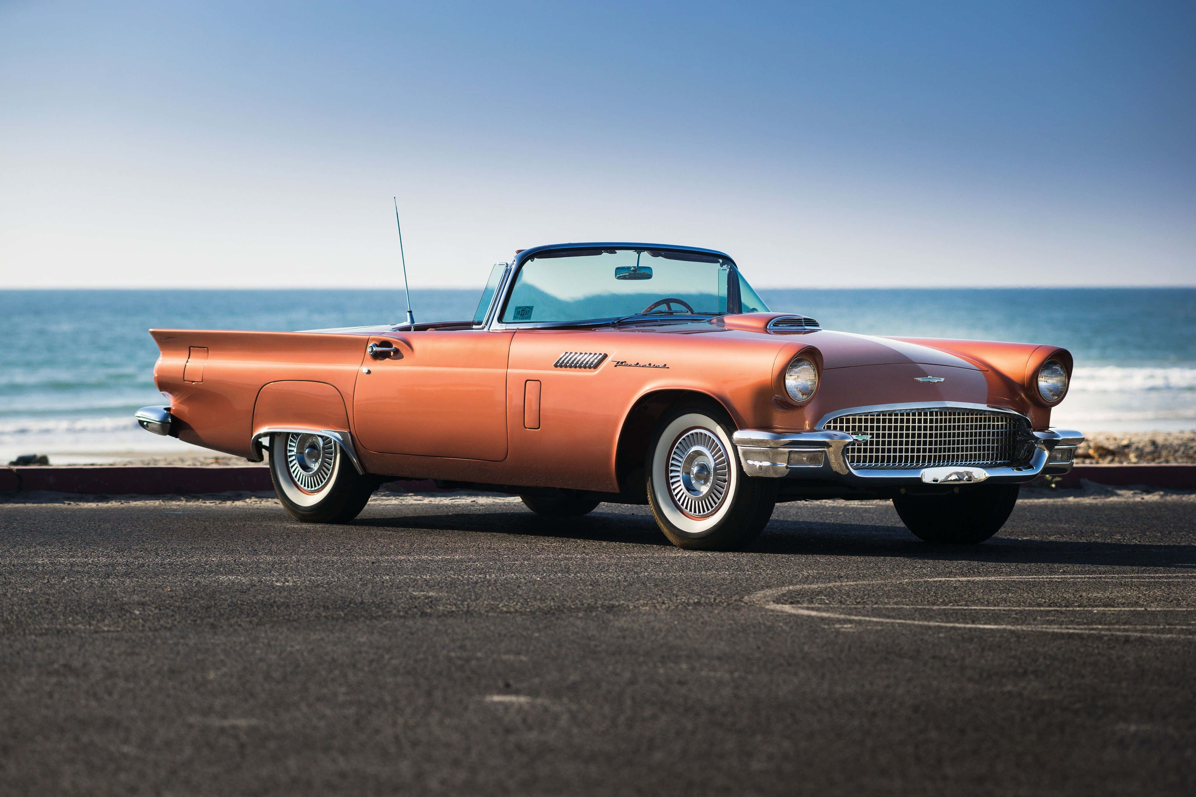 Ford Thunderbird 桌布 壁紙 — 一輛復古橙色開篷車停泊在沿海公路上，背後有海浪拍打，天空清澈湛藍。