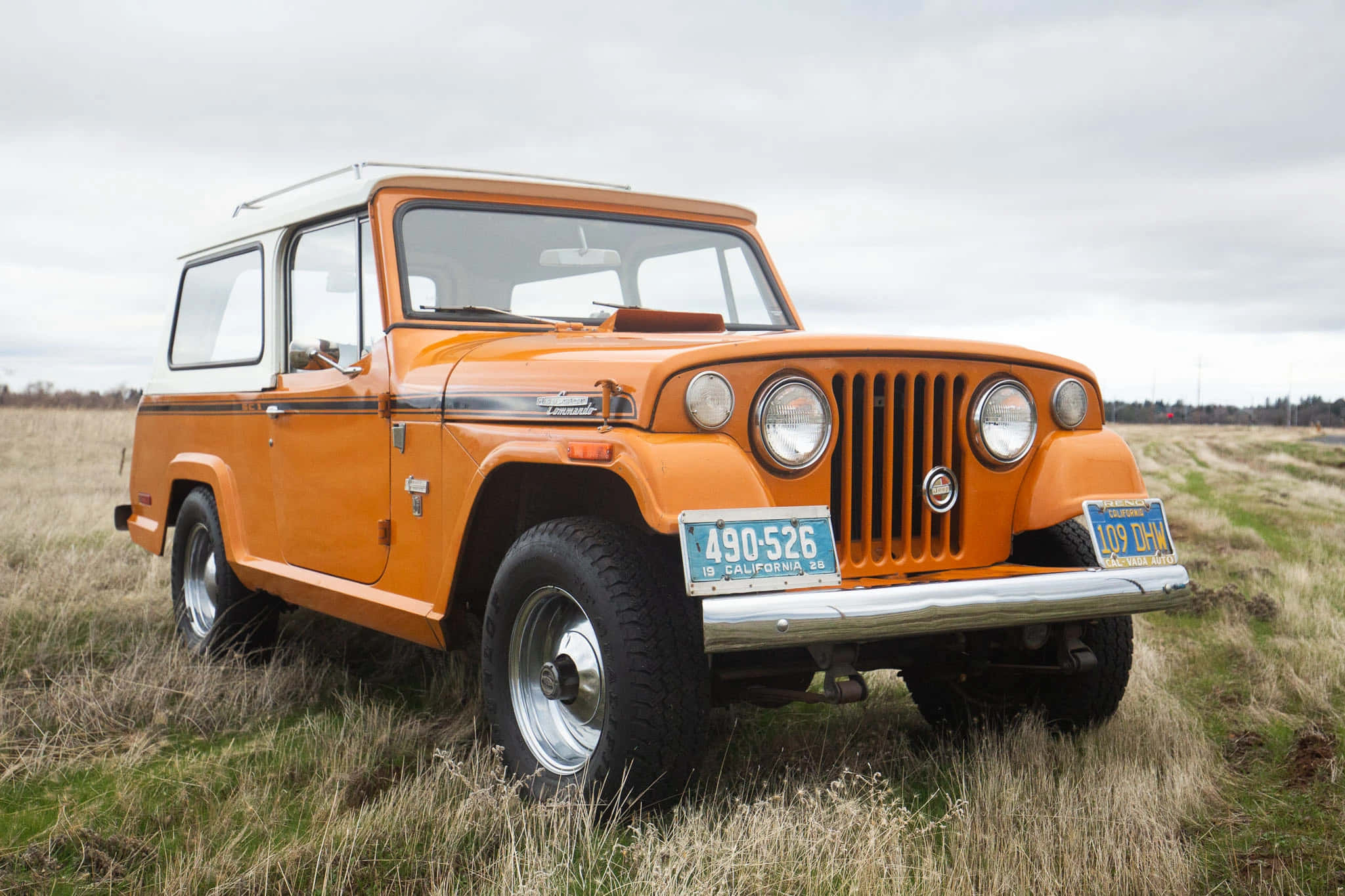 Jeepster 桌布 壁紙 — 一輛復古橙色Jeep Wagoneer，白色車頂，停泊在草地田野上，天空多雲。