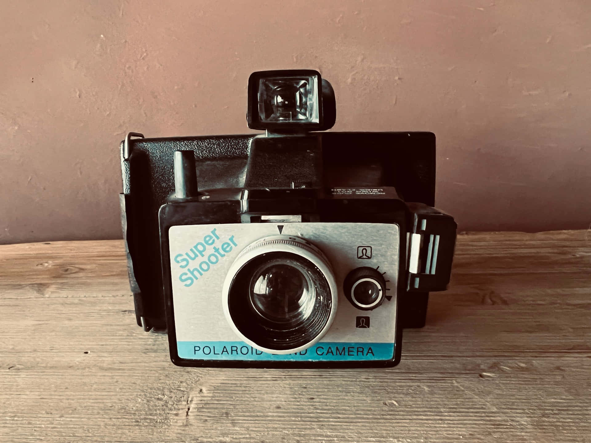  壁紙 — 一部復古Polaroid Super Shooter相機放置在經風化的木質表面上，背景是柔和的粉紅色牆壁。