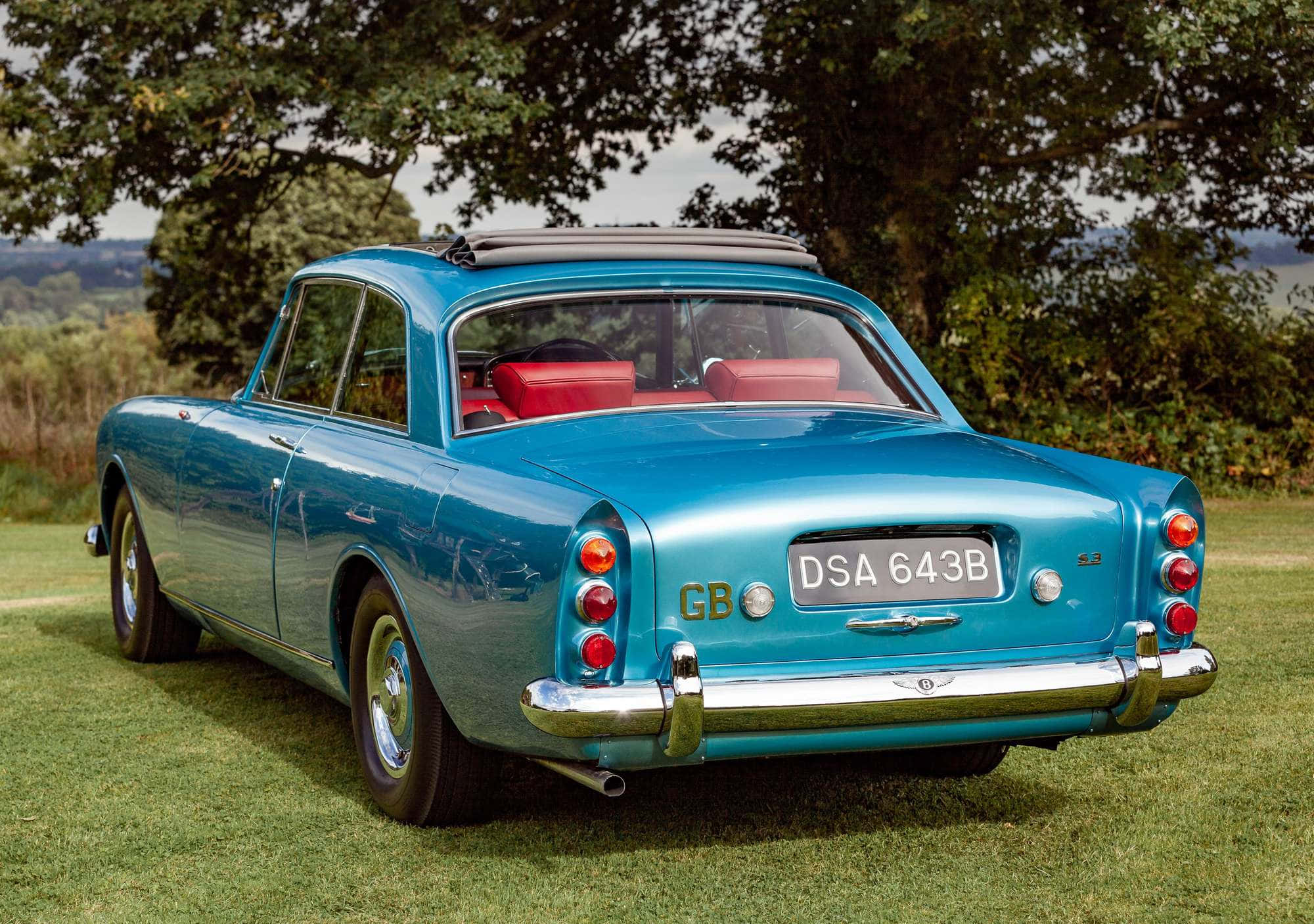 Bentley S3 桌布 壁紙 — 一輛復古青綠色Bentley S3，紅色內飾，停在樹葉茂密的樹下的草地上。