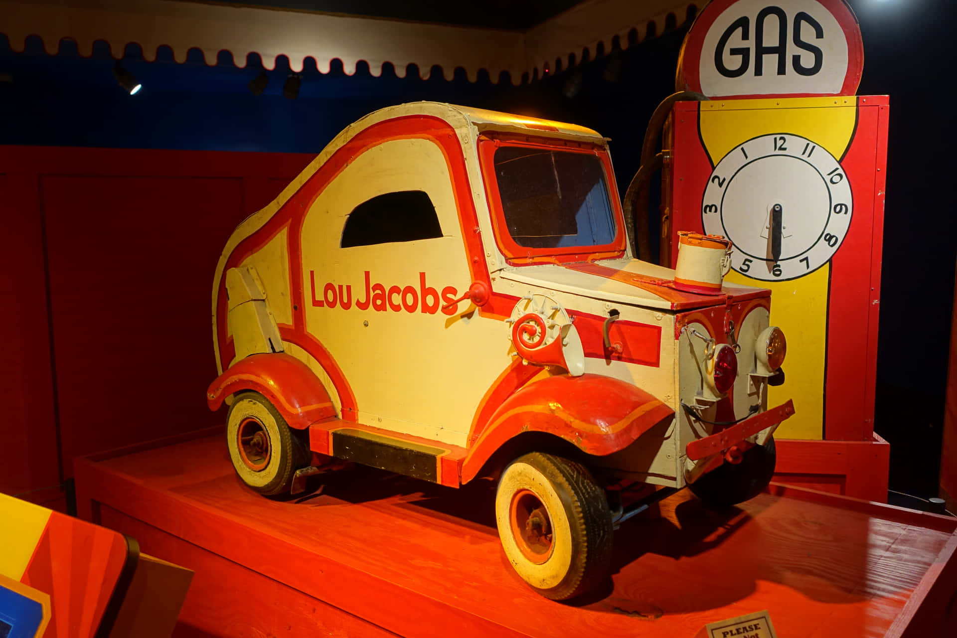  壁紙 — 一輛標有 Lou Jacobs 的復古紅白玩具車，在溫暖的博物館燈光下，停放在一台復古油泵旁。