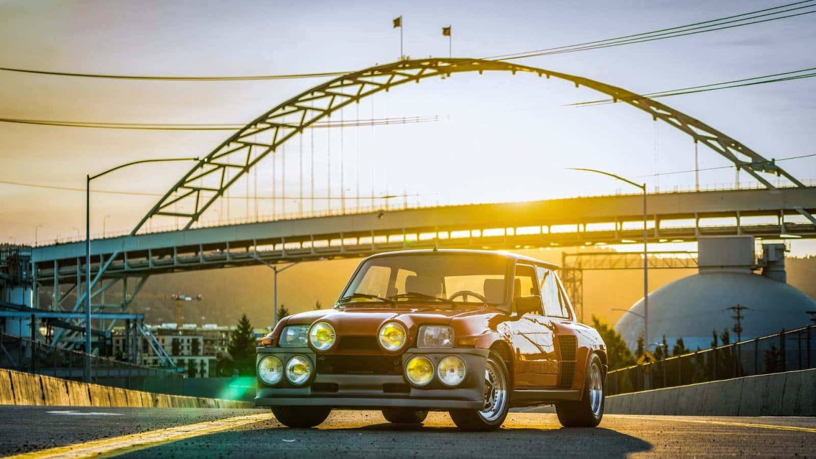 Renault 5 Turbo 桌布 壁紙 — 一輛復古紅色BMW 3.0 CSL配備四盞頭燈，在金色日落時分停泊在鋼拱橋下。