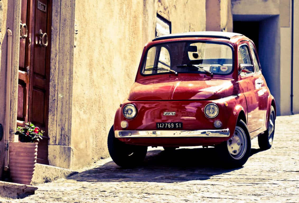 Fiat 500 桌布 壁紙 — 一架復古紅色Fiat 500停泊在陽光普照的鵝卵石街道上，旁邊是一棟有盆栽花卉的風化建築。