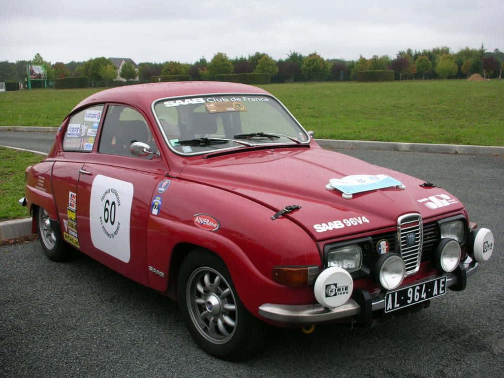 Saab 96 桌布 壁紙 — 一架復古紅色薩博拉力賽車，編號60，停泊在瀝青路上，靠近草地，天空多雲。