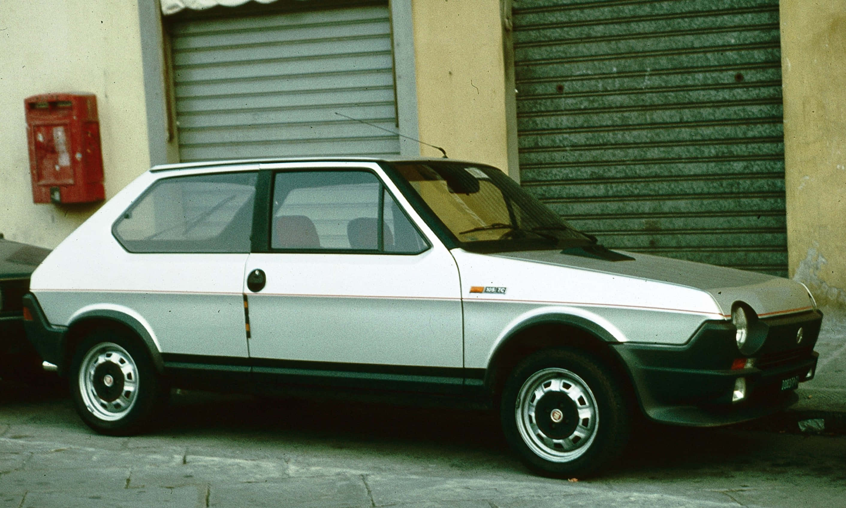 Fiat Ritmo 桌布 壁紙 — 一輛復古銀白色快意 Panda 停泊在鵝卵石街道上，旁邊有一個紅色郵箱和金屬百葉窗。