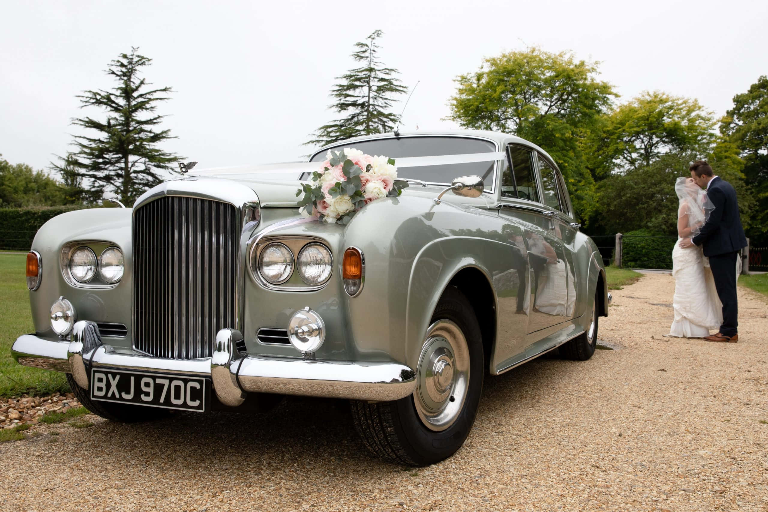 Bentley S3 桌布 壁紙 — 一輛裝飾著花朵的復古銀色賓利婚禮車停在碎石上，新娘和新郎在附近接吻。