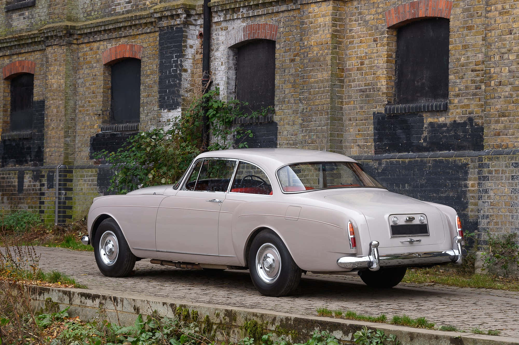 Bentley S2 桌布 壁紙 — 一輛復古銀色跑車停泊在鵝卵石小徑上，旁邊是一座風化的磚砌建築，長滿了茂密的樹葉。