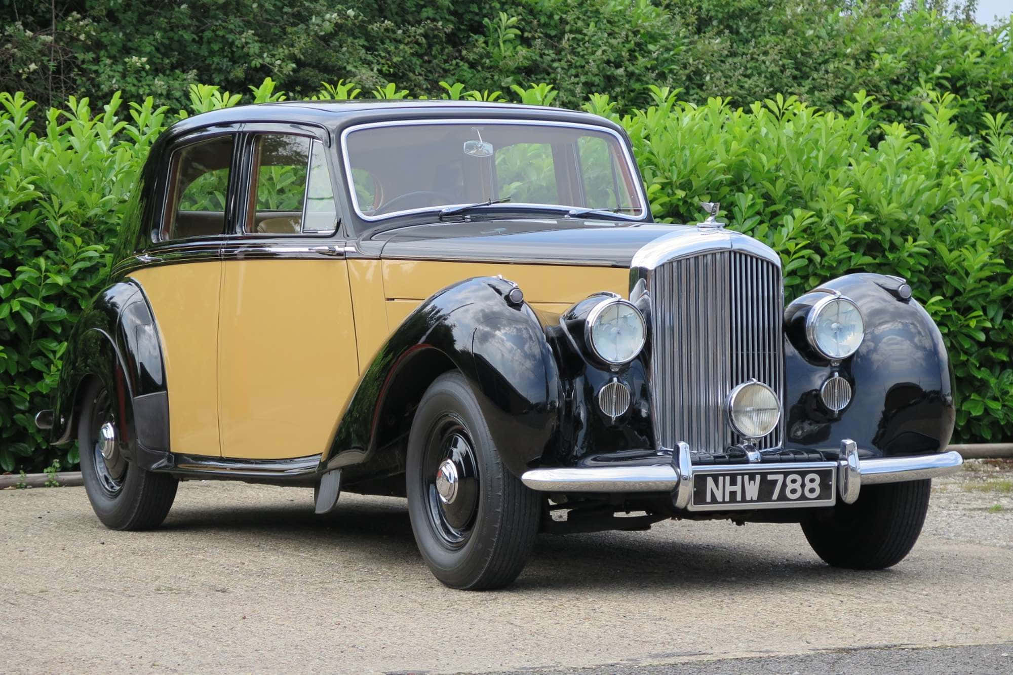 Bentley Mark Vi 桌布 壁紙 — 一輛復古雙色汽車，黑色和芥末黃色車漆在日光下閃閃發光，停在碎石路上，旁邊是茂密的綠色樹籬。