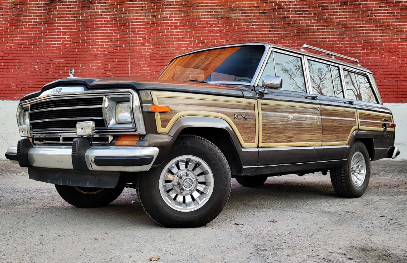 Jeep Wagoneer 桌布 壁紙 — 一輛復古木紋車身的Jeep Grand Wagoneer停泊在紅磚牆前的碎石地面上。