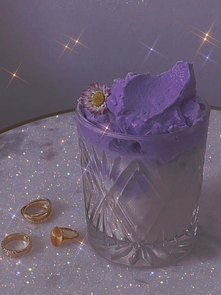欣賞Violet Aesthetic的魔幻之美 壁紙 — 一杯紫色雪糕，上面放著一朵花，放在閃閃發光的表面上，附近有閃光和金色戒指。