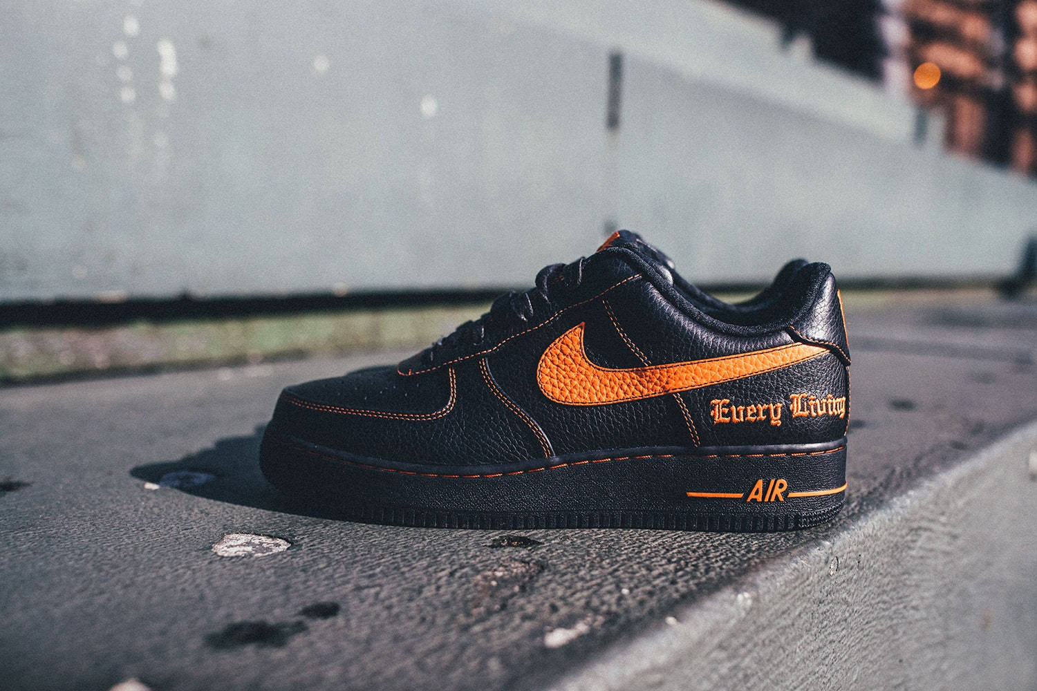 VLONE x Nike Air Force One 鞋款 壁紙 — 一雙黑色Nike Air Force 1運動鞋，帶有鮮豔的橙色點綴和「Every Living」字樣，在溫暖的陽光下放置於粗糙的城市人行道上。