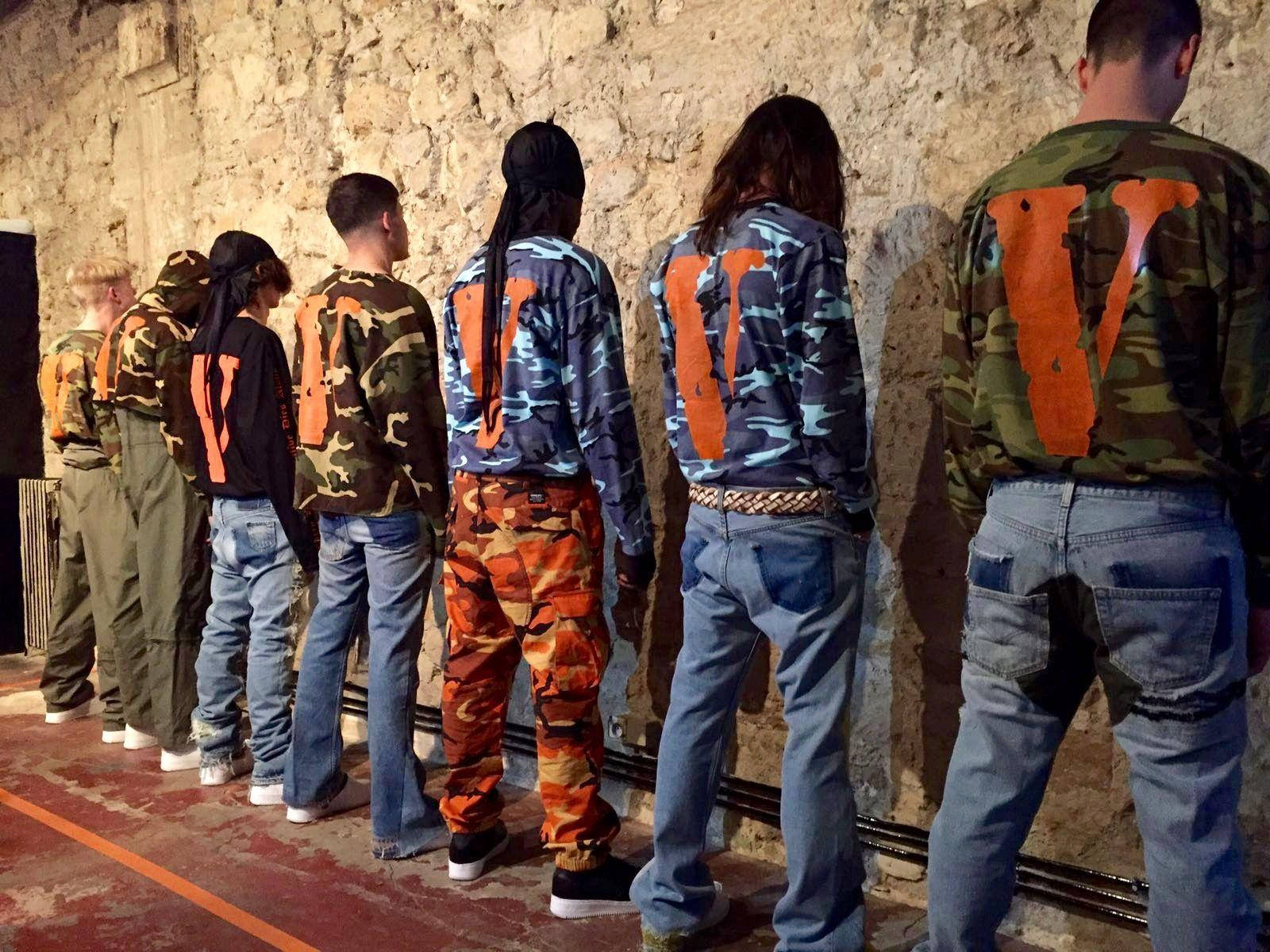 VLONE 模特面對牆壁 壁紙 — 一群穿著迷彩外套、帶有橙色「V」標誌的人，排成一排站在粗糙的石牆前。