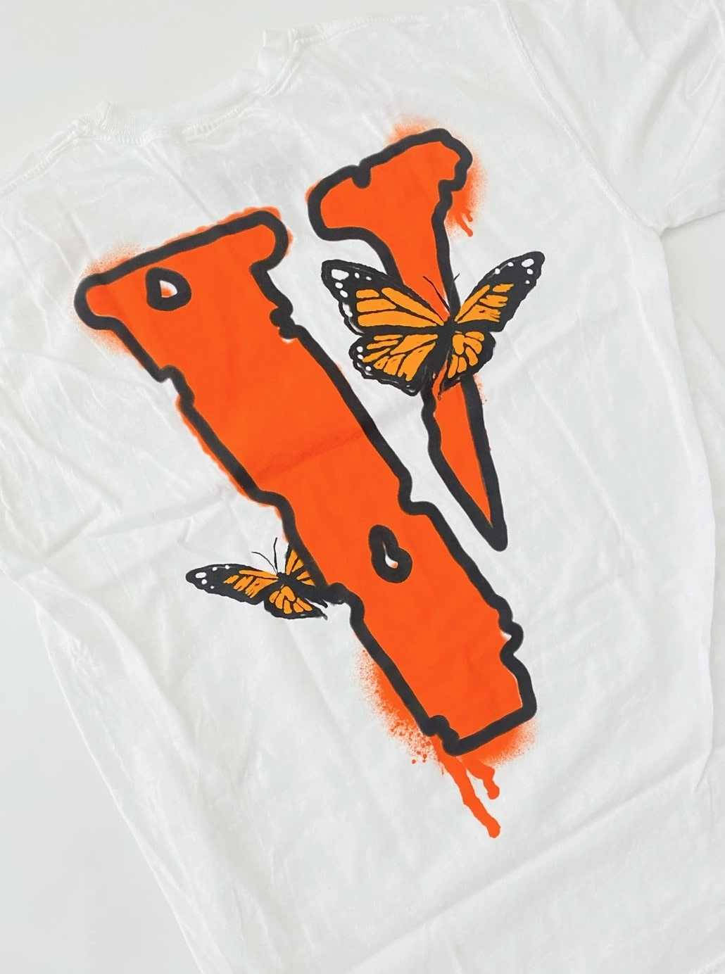 VLONE 橙色蝴蝶 壁紙 — 一件白色T恤上有一個大膽的橙色噴漆V字，兩隻帝王斑蝶棲息在它的曲線上。