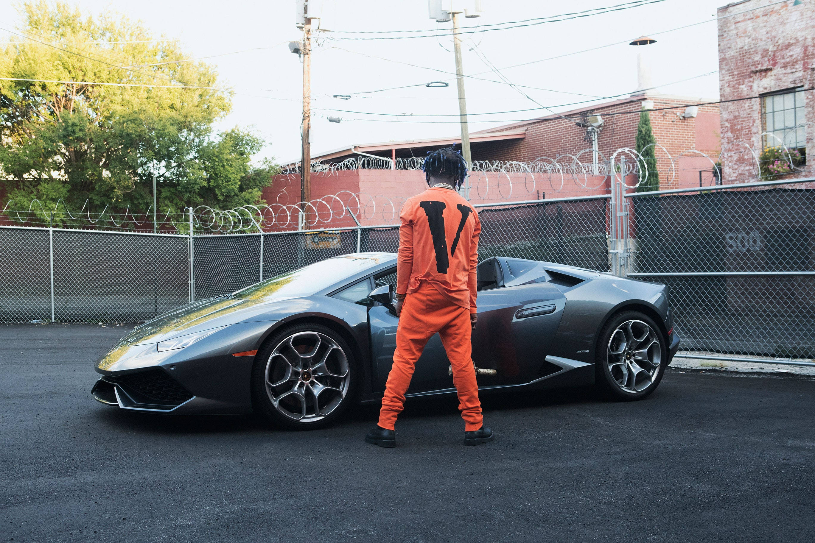 VLONE 橙色連帽衫 壁紙 — 一名身穿橙色連身衣的人站在一個有鐵絲網圍欄和磚砌建築的市區空地，旁邊是一輛時尚的灰色林寶堅尼。
