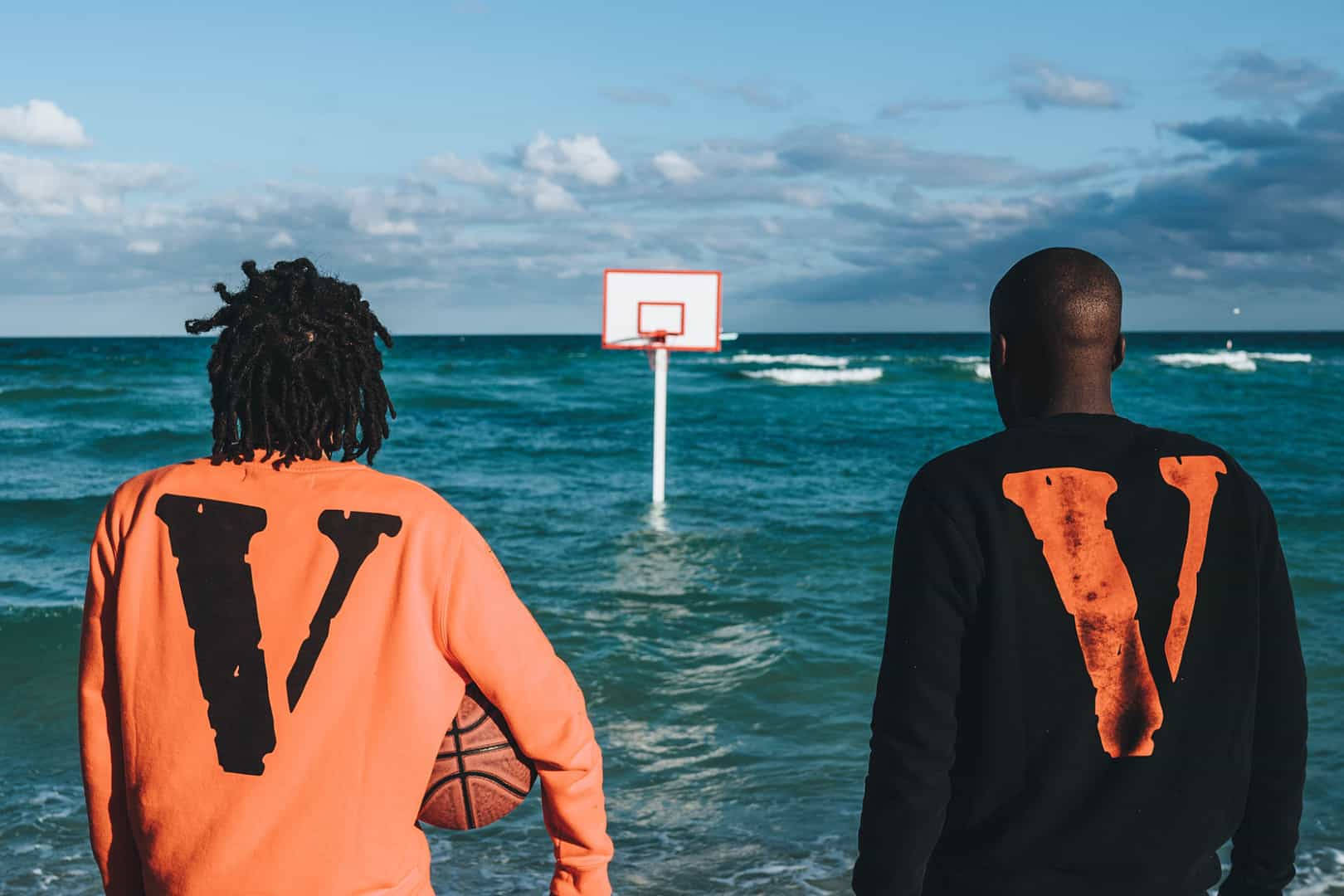 VLONE 海灘背景廣告 壁紙 — 兩名男子穿著相配的「V」連帽衫，站在齊腰深的海水中，面對著一個部分多雲的天空下浸沒的籃球框。