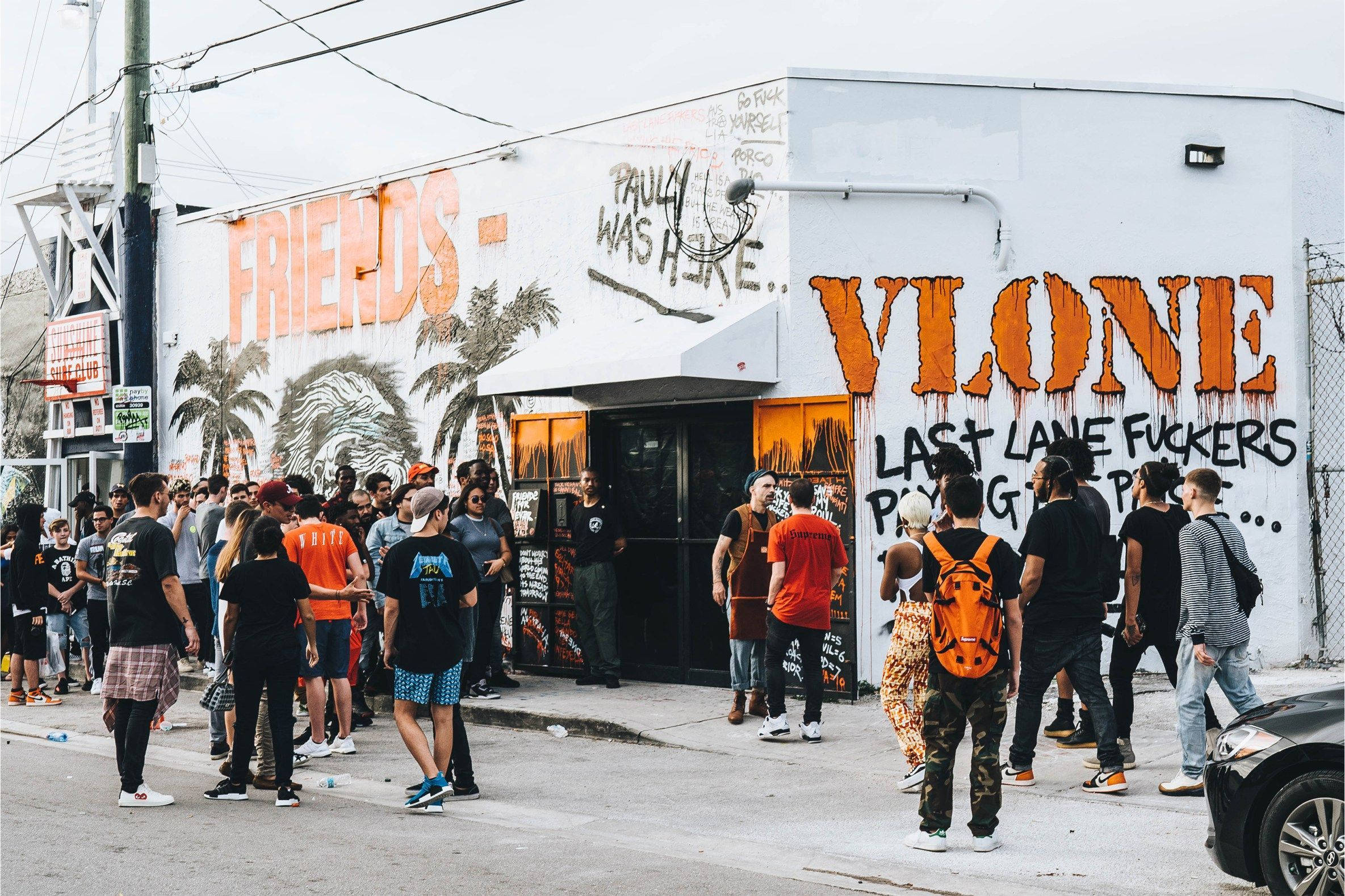 VLONE 街頭藝術 壁紙 — 人群聚集在一棟被塗鴉覆蓋的建築物外，建築物上有大膽的橙色和黑色街頭藝術，天空多雲。