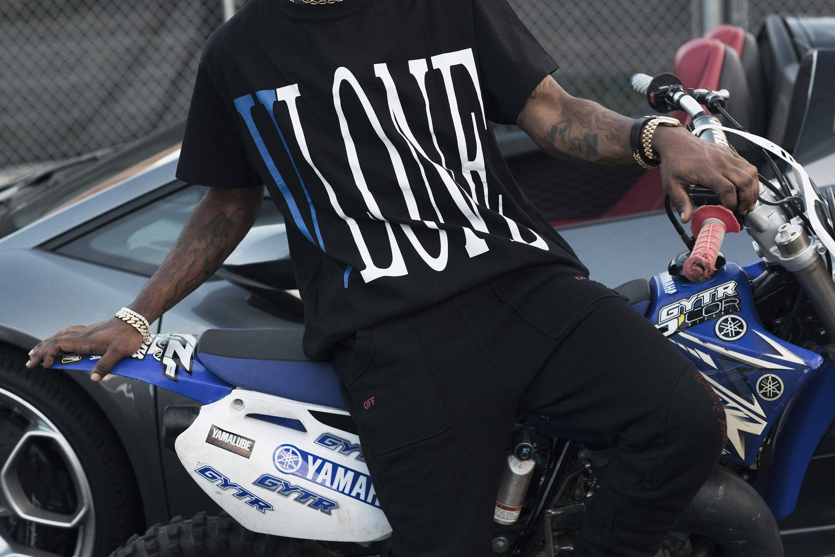VLONE 黑色上衣模特照 壁紙 — 一個有紋身的人坐在藍白相間的Yamaha越野電單車旁邊，旁邊是一輛時尚的灰色跑車。
