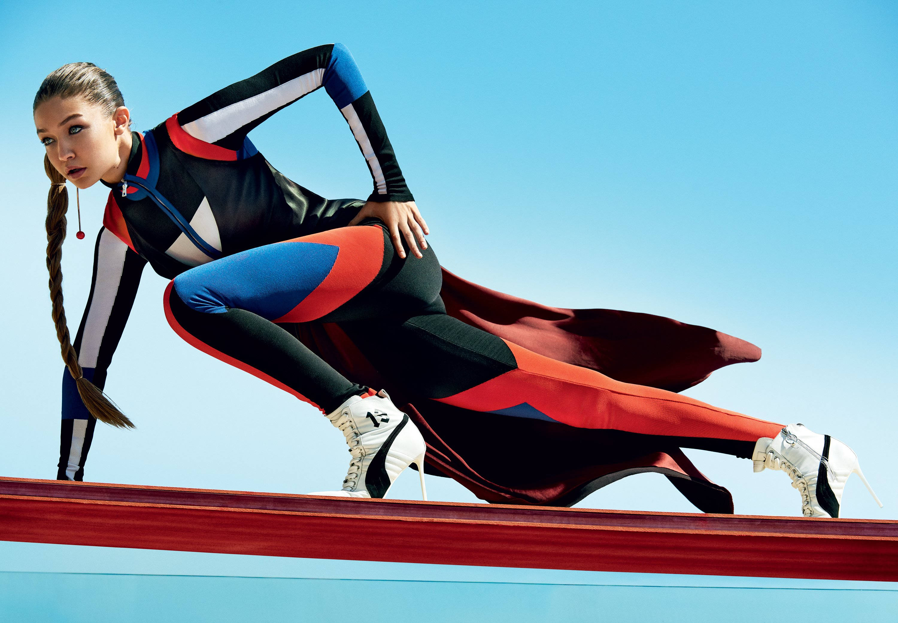 吉吉·哈迪德 運動風《Vogue》封面 壁紙 — 一位女性速度滑冰選手穿著大膽的紅、藍、黑色緊身衣，在紅色跑道上向前傾，背景是晴朗的藍天。