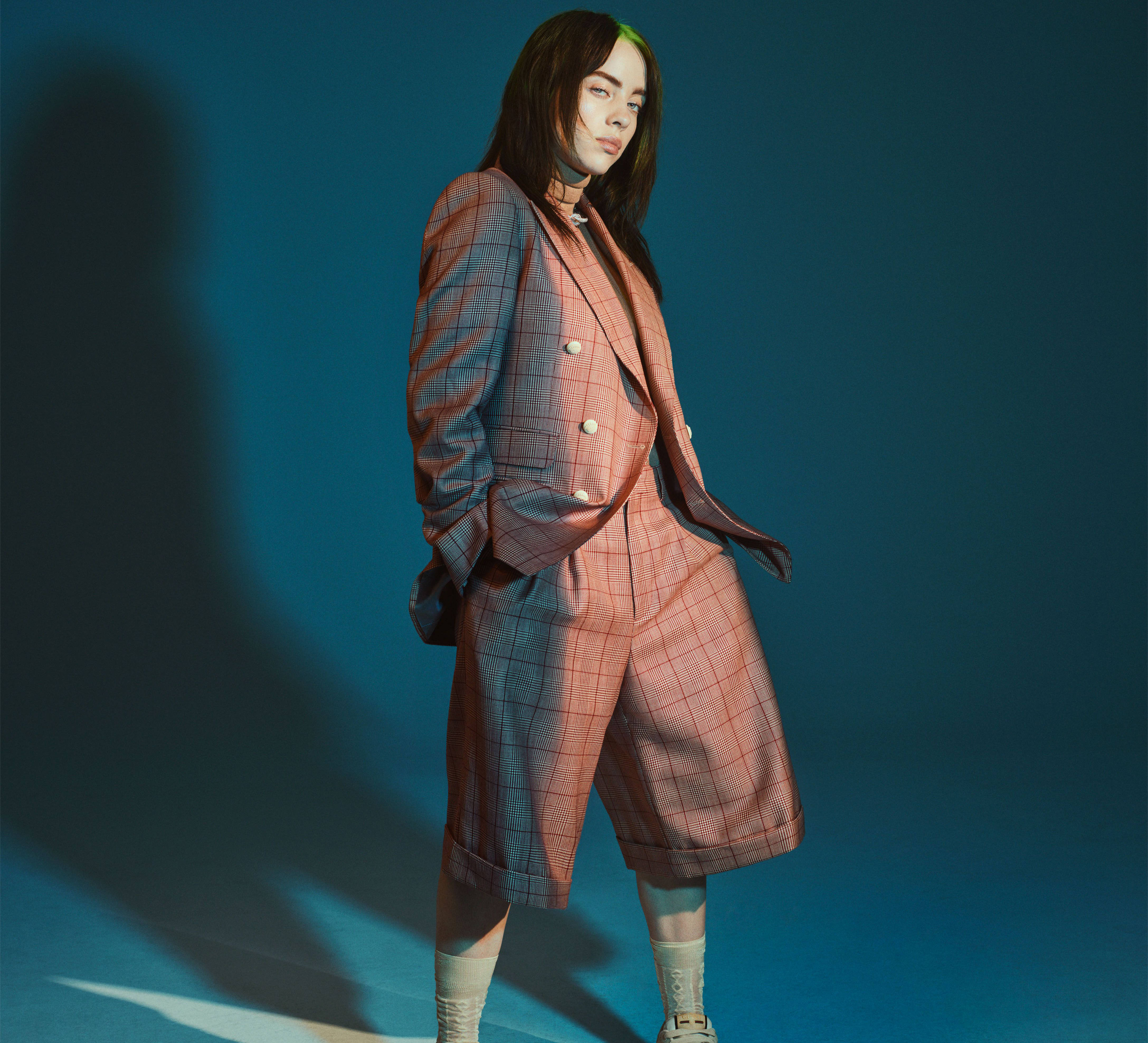 《Vogue》比莉·艾利什 4K 壁紙 — 一個人穿著粉紅色格紋西裝，站在深藍色工作室背景前，戲劇性的燈光投射出長長的影子。