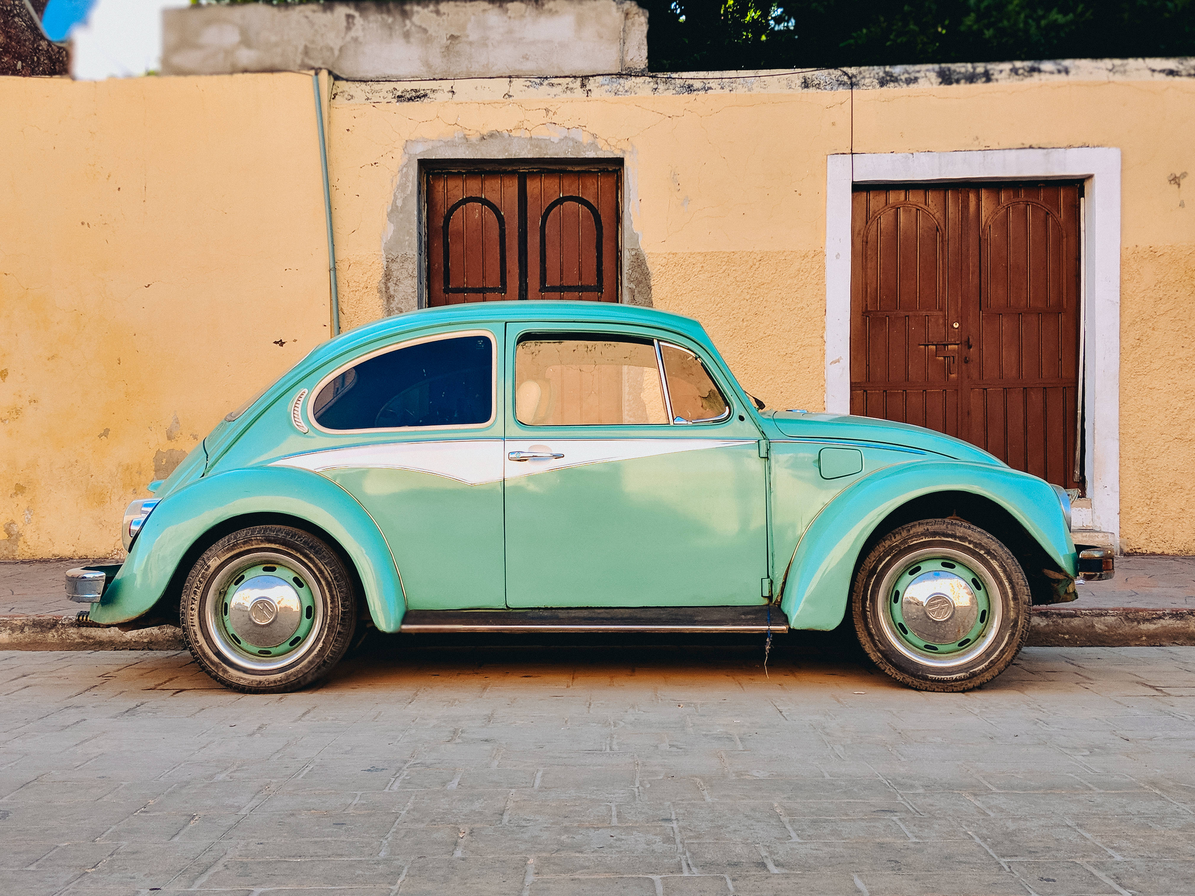 復古 Volkswagen Beetle 壁紙 — 一輛復古藍綠色福士甲蟲車停泊在陽光明媚的鵝卵石街道上，旁邊是帶有木門的風化黃色牆壁。