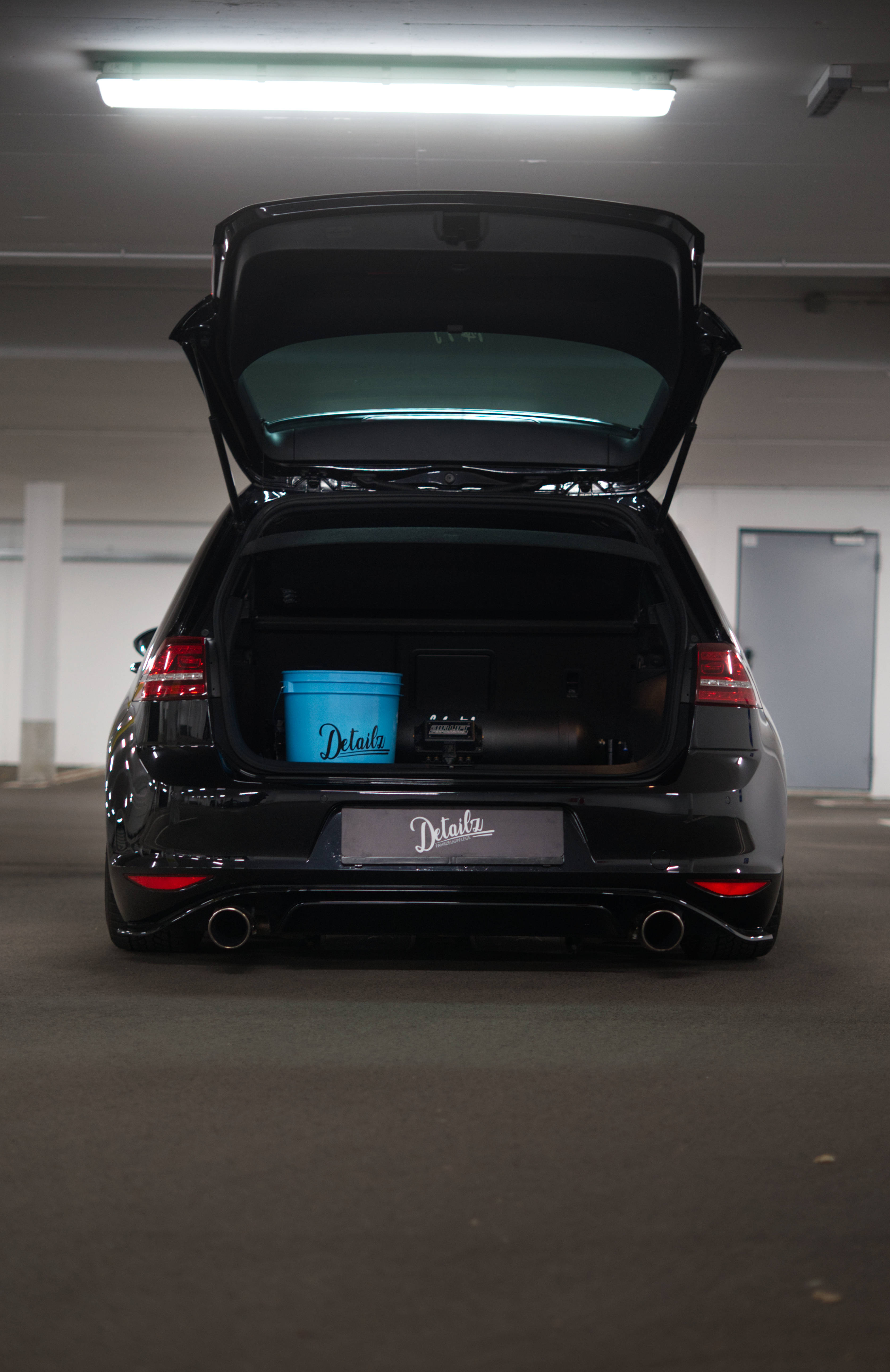 Volkswagen Golf GTI 開尾箱 壁紙 — 一輛亮黑色旅行車，尾門打開，露出車內一個藍色細節清潔桶，停在光線昏暗的車庫中。