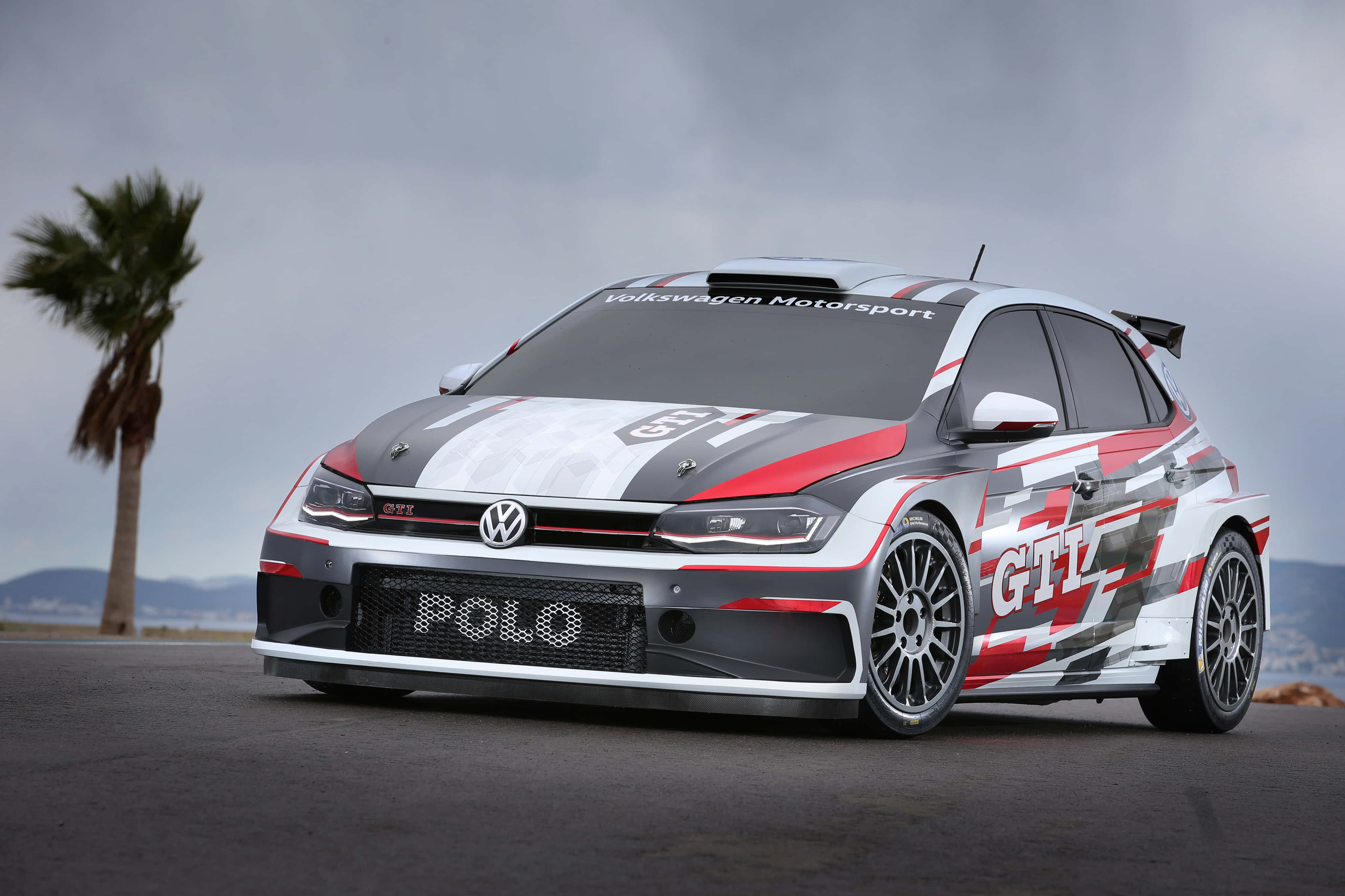 Volkswagen Polo 桌布 壁紙 — 一輛紅白黑塗裝的福士Polo GTI拉力賽車停在柏油路上，旁邊有棕櫚樹，天空多雲。