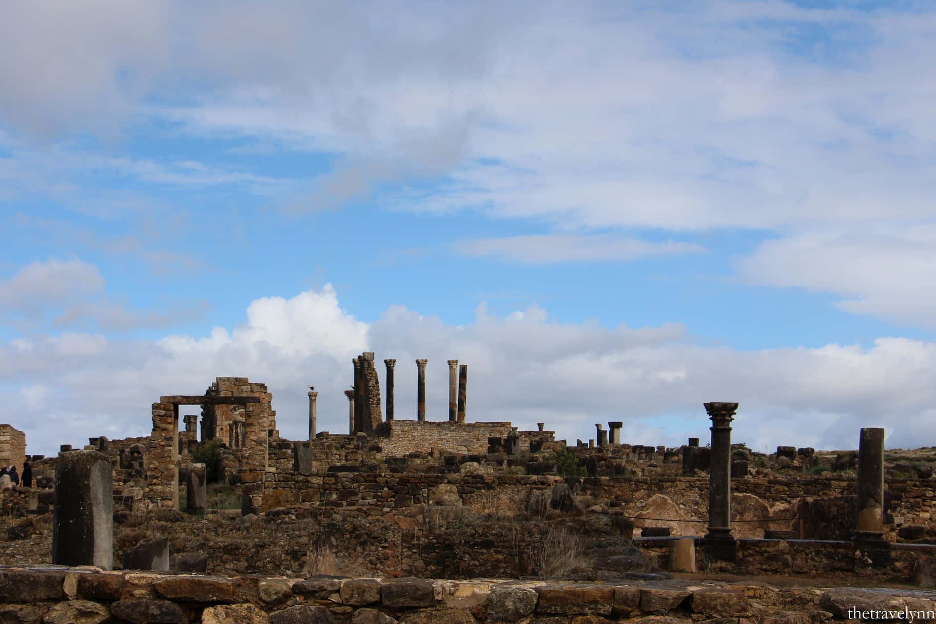 Volubilis 上空雲層 壁紙 — 古代石頭廢墟，斷裂的柱子，矗立在明亮的藍天下，散落著白雲。