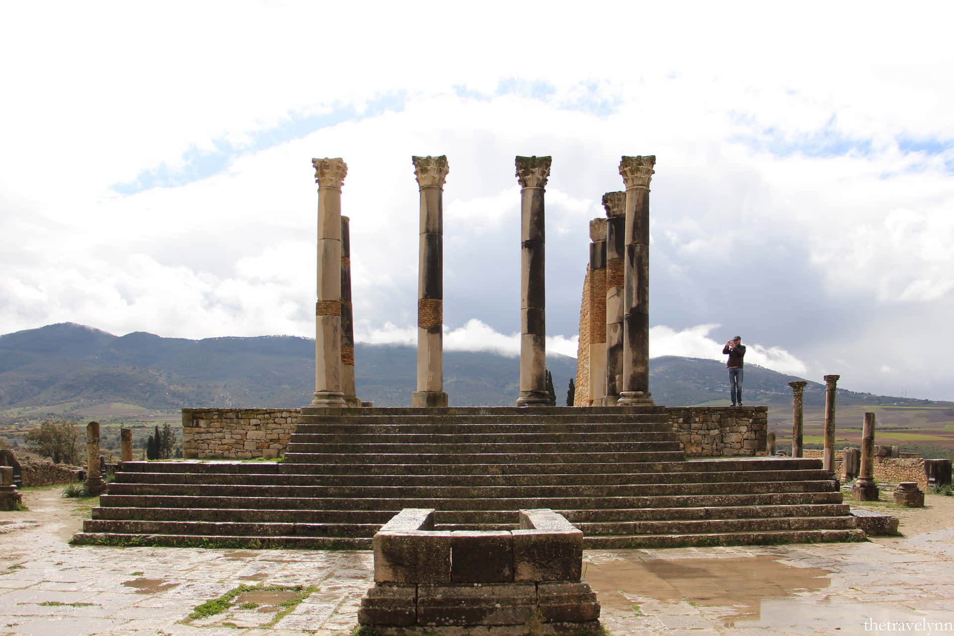 Volubilis 的基座與建築殘跡 壁紙 — 古老石廟遺跡，高柱與寬階聳立於多雲天空與遠山之前。