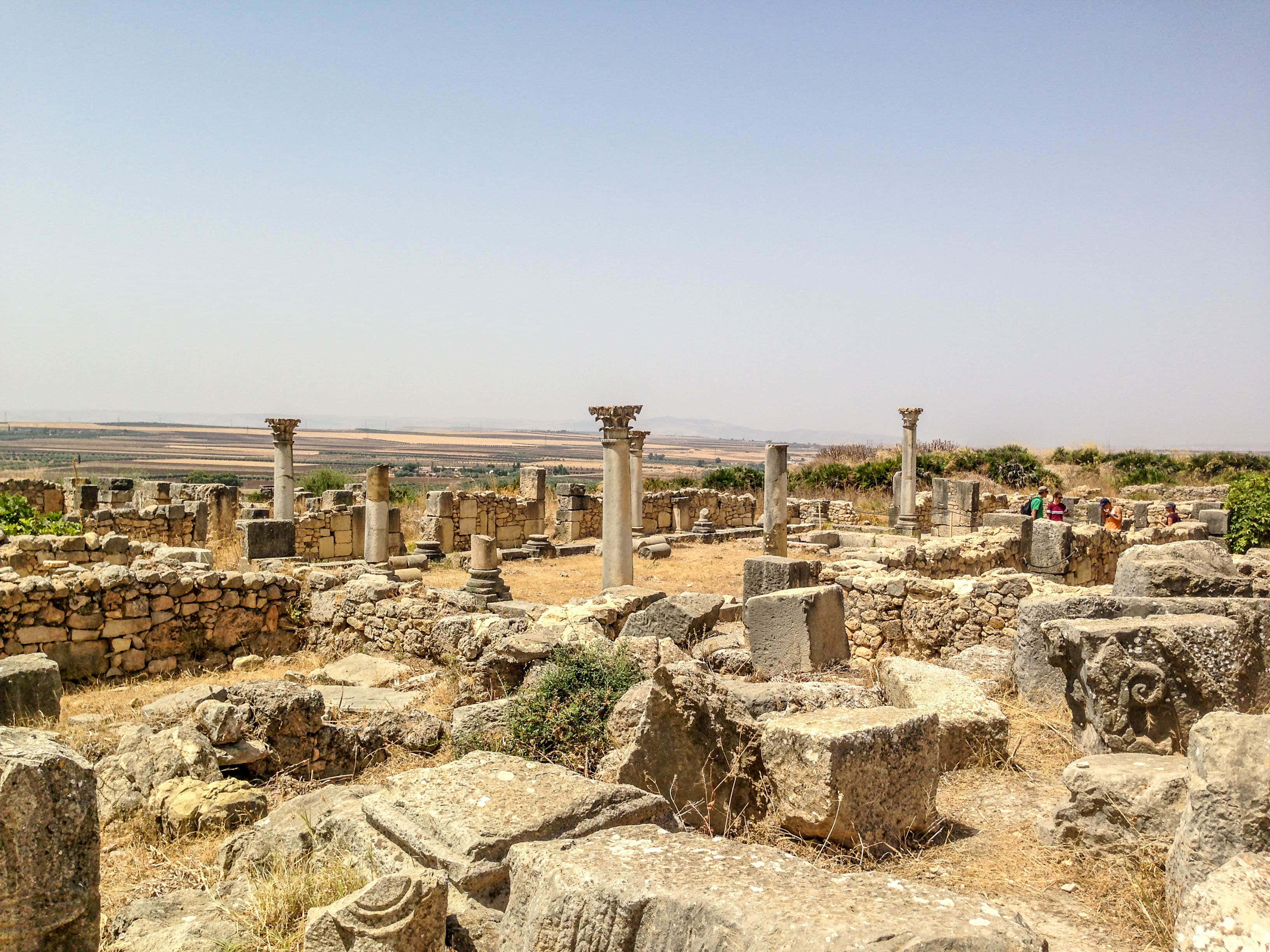 Volubilis 的巨大石塊 壁紙 — 古代石頭遺跡，有豎立的柱子和散落的石塊，在晴朗的藍天下，遠處有山丘。
